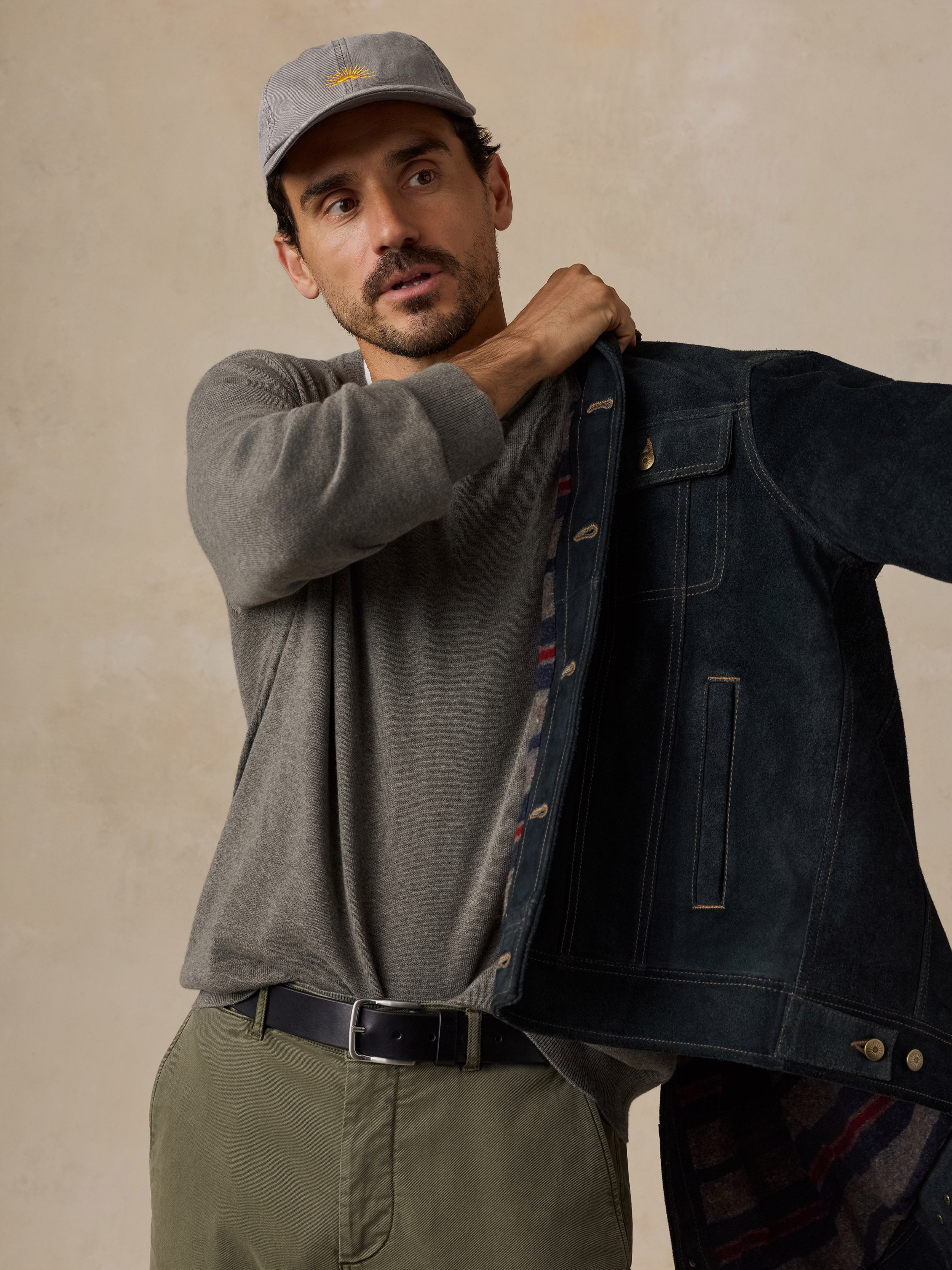 A man wearing a green knit sweater, olive pants, a gray cap, and a dark suede jacket.