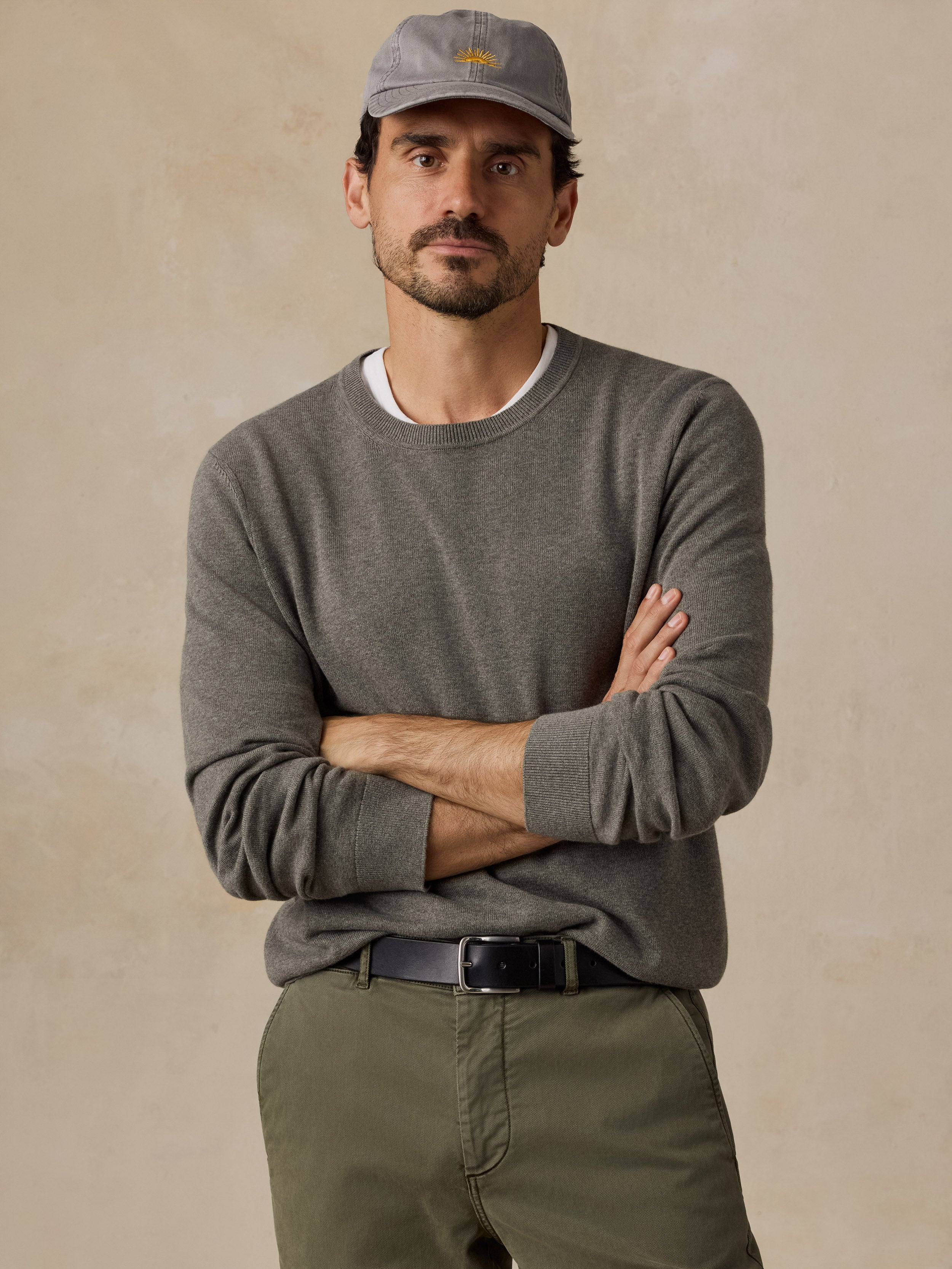 A man wearing a green crewneck sweater, olive green pants, a gray cap, and a black belt.
