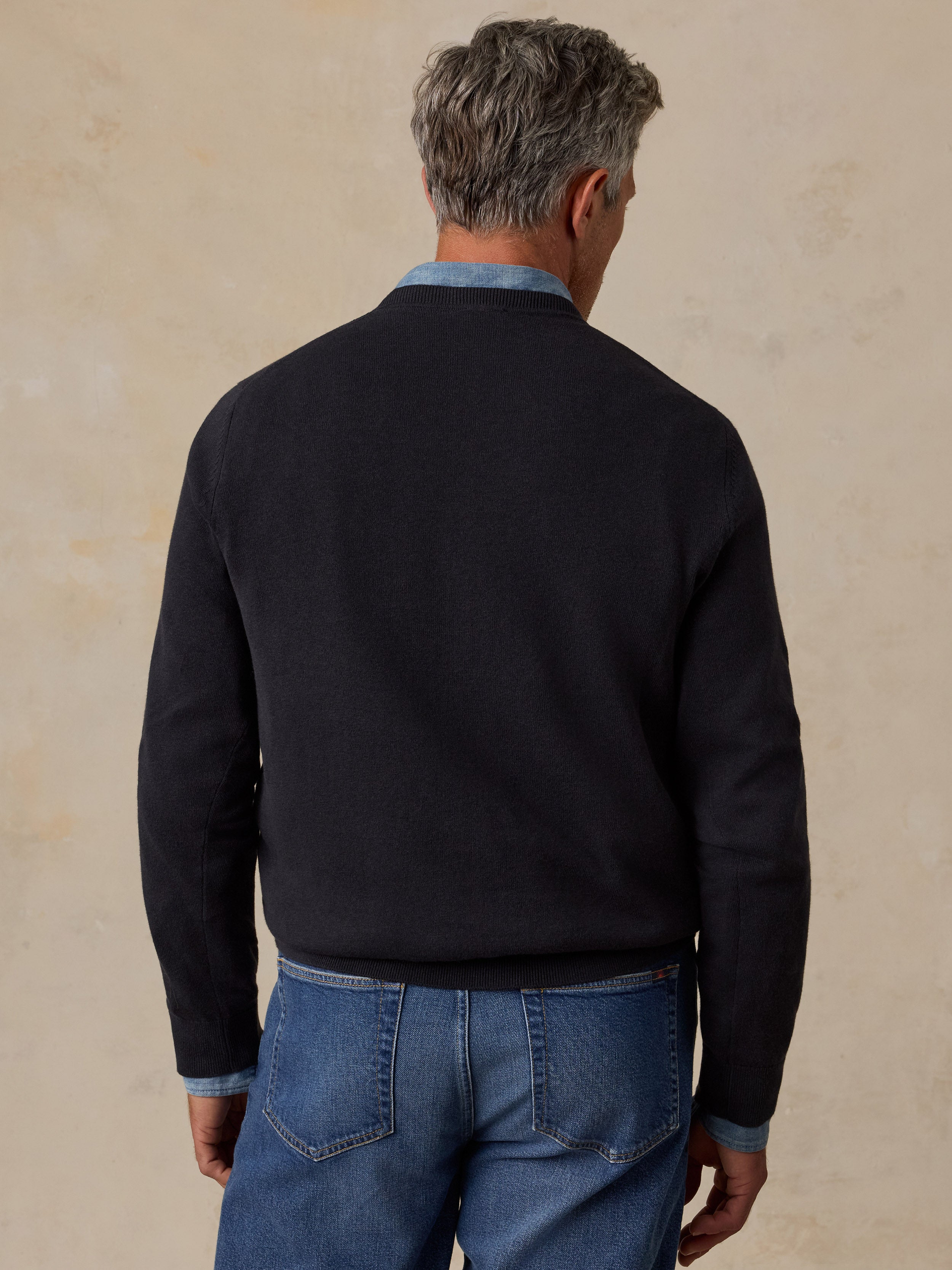 A man viewed from behind wearing a dark navy knit sweater over a light blue collared shirt and blue jeans.