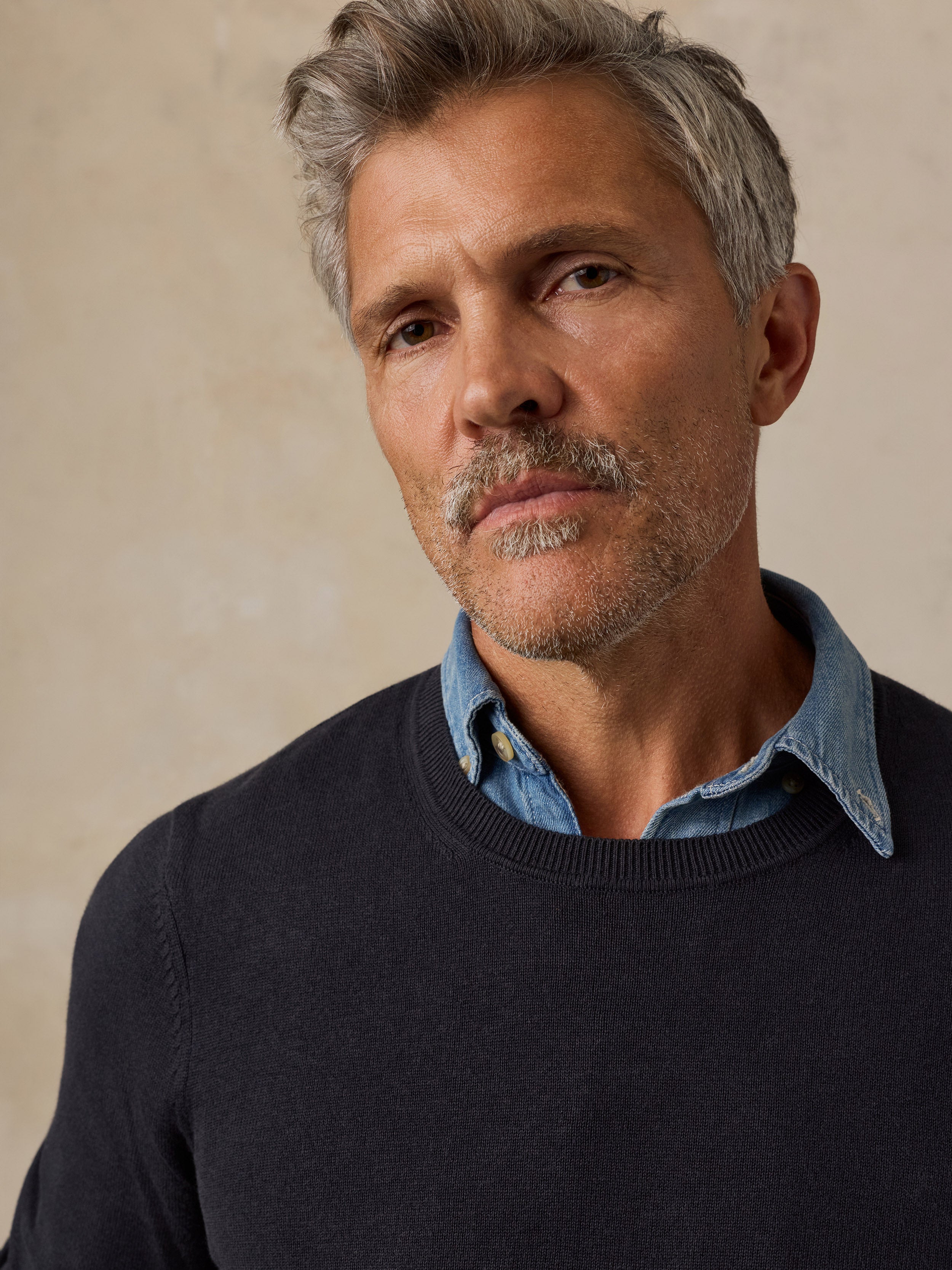 A close-up of a man wearing a dark navy crewneck sweater over a light blue denim shirt.