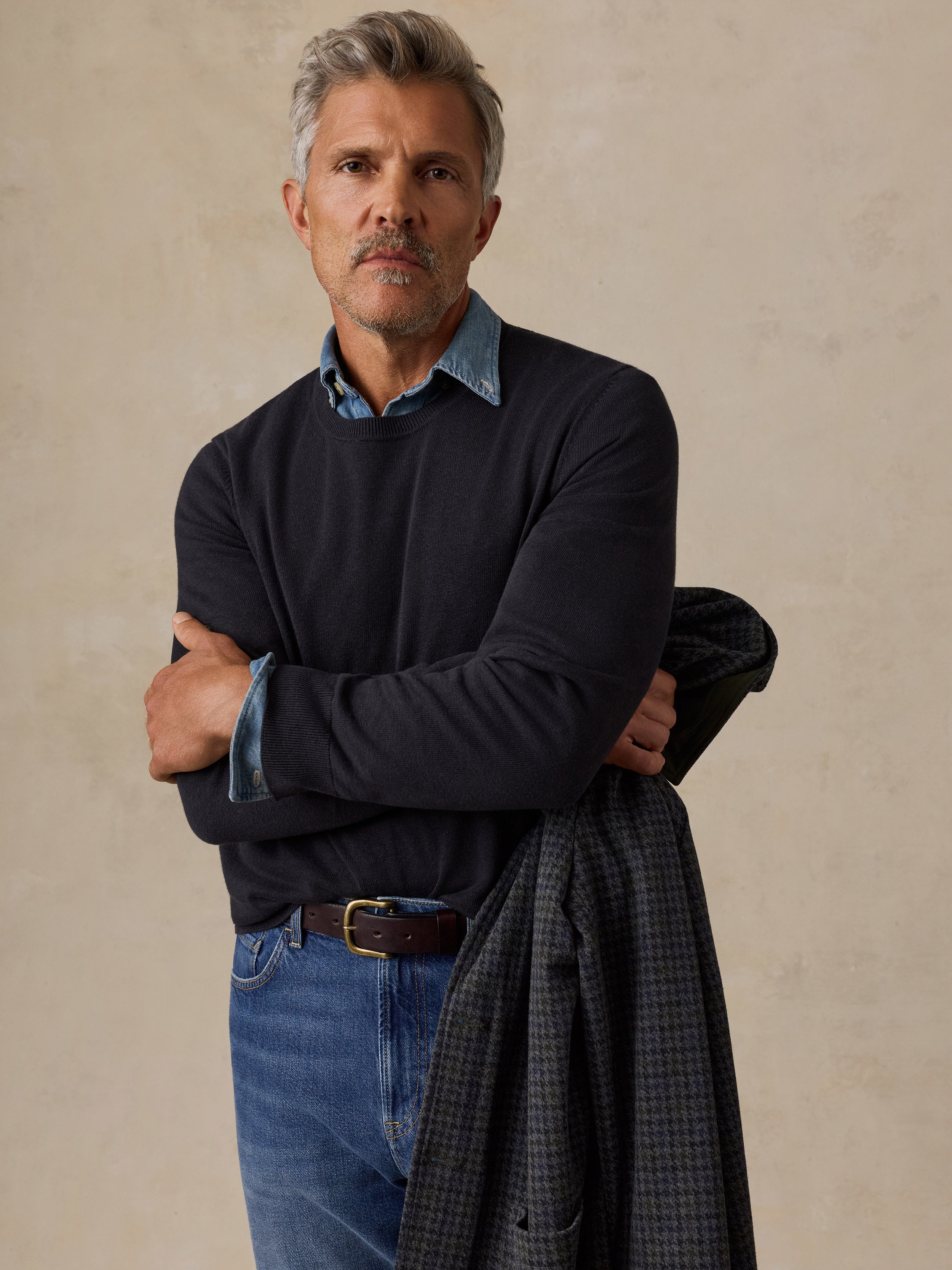 A man wearing a dark navy long-sleeve sweater over a light blue collared shirt, and blue jeans.