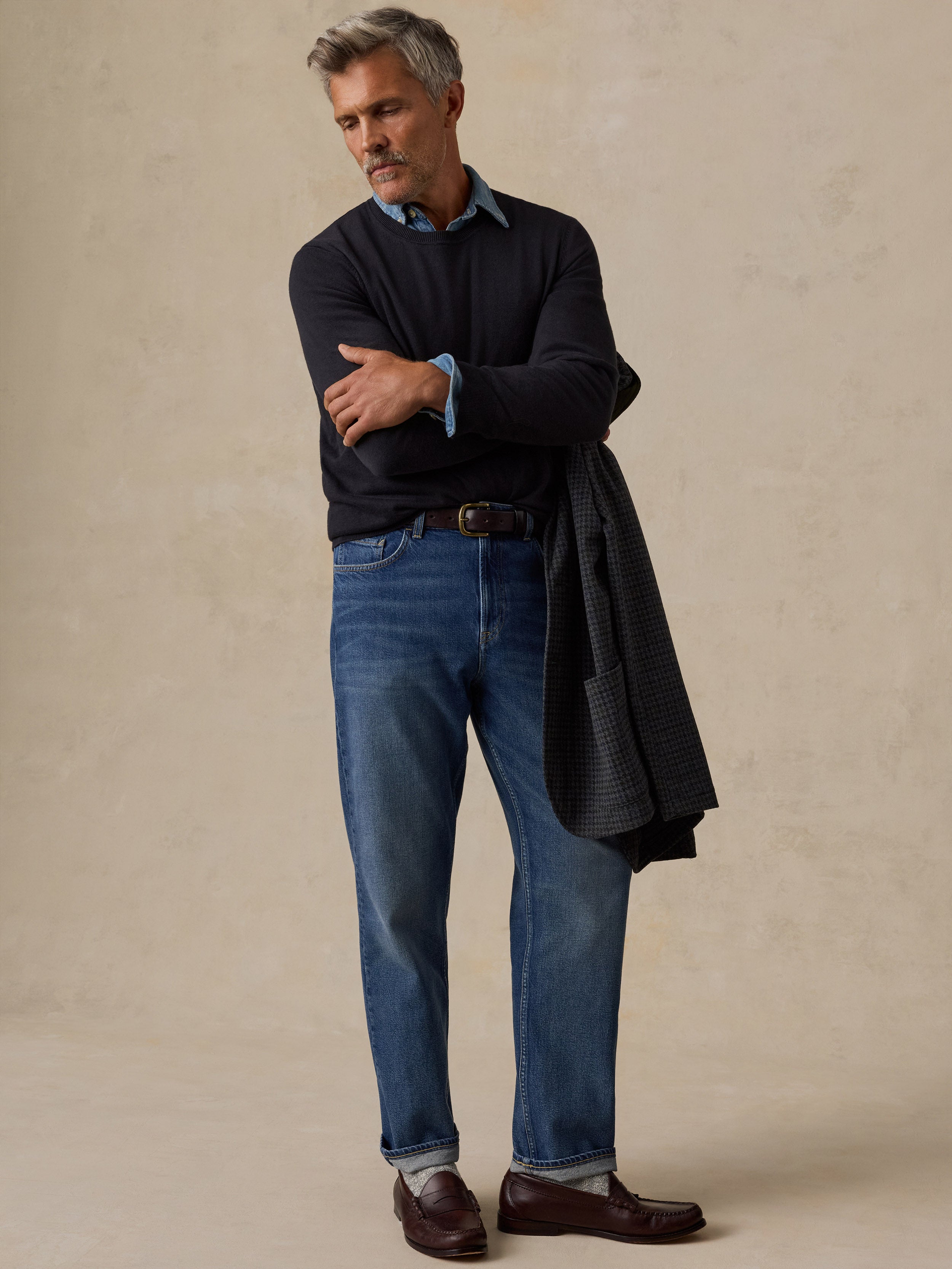 A man wearing a long-sleeve sweater over a light blue collared shirt, blue jeans, and brown loafers.