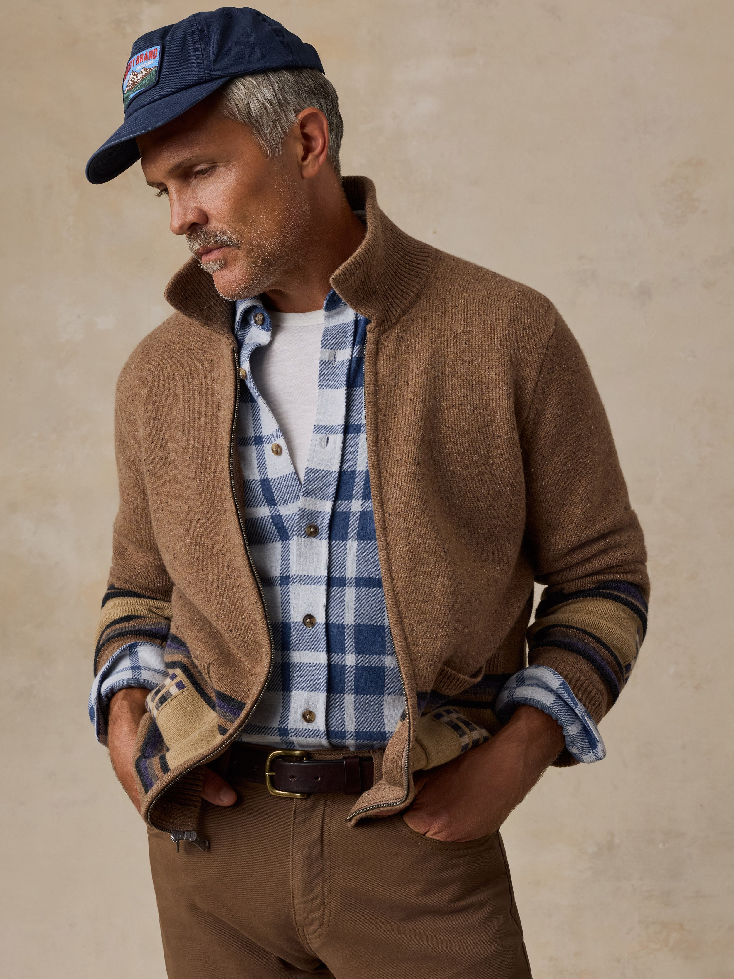 A man wearing a brown knit zip-front jacket over a blue and white plaid button-down shirt, paired with brown pants and a navy baseball cap, posed against a textured beige background.