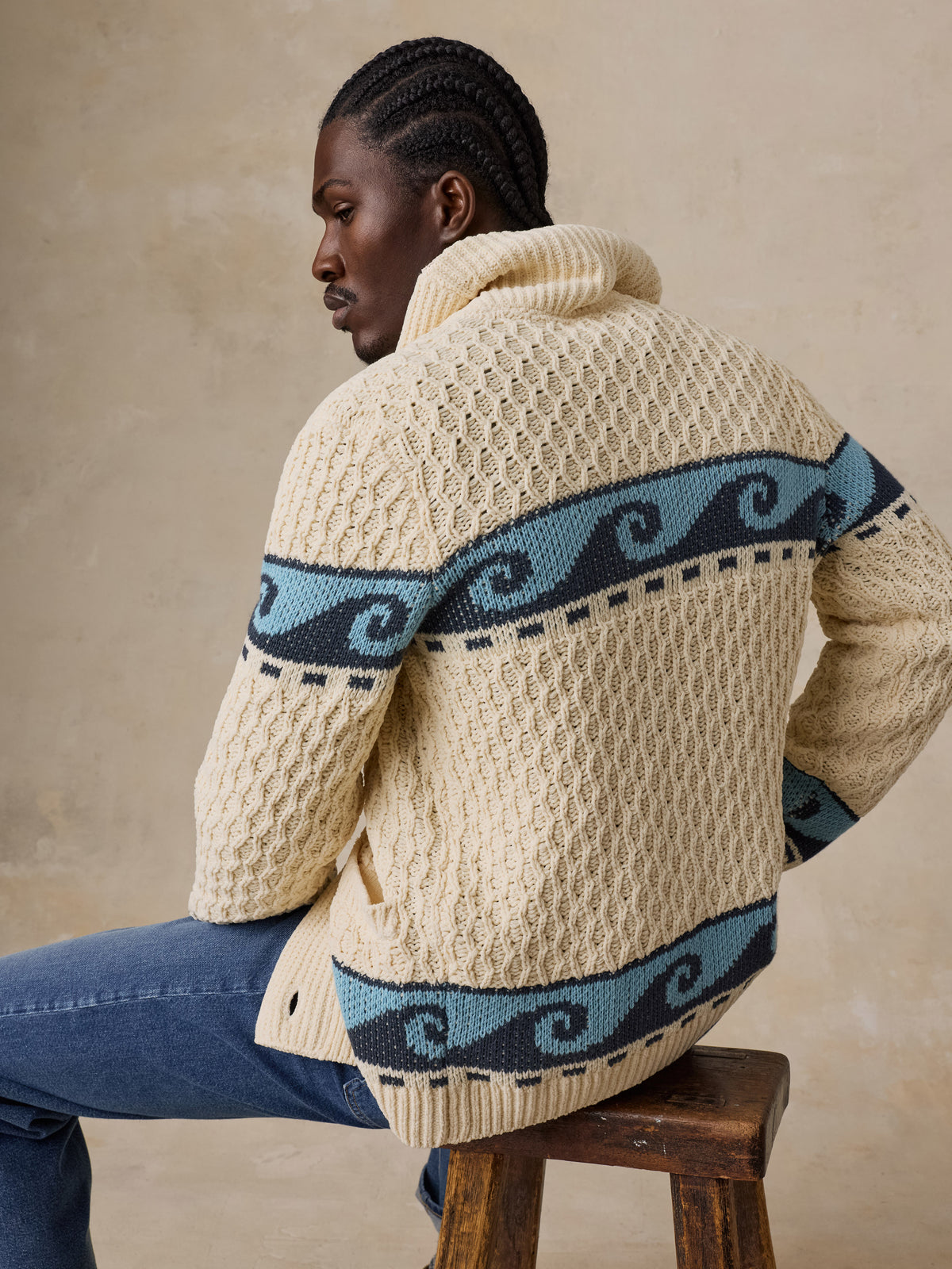 A man viewed from behind wearing a cream and blue printed cardigan with blue jeans, image 2 of 7
