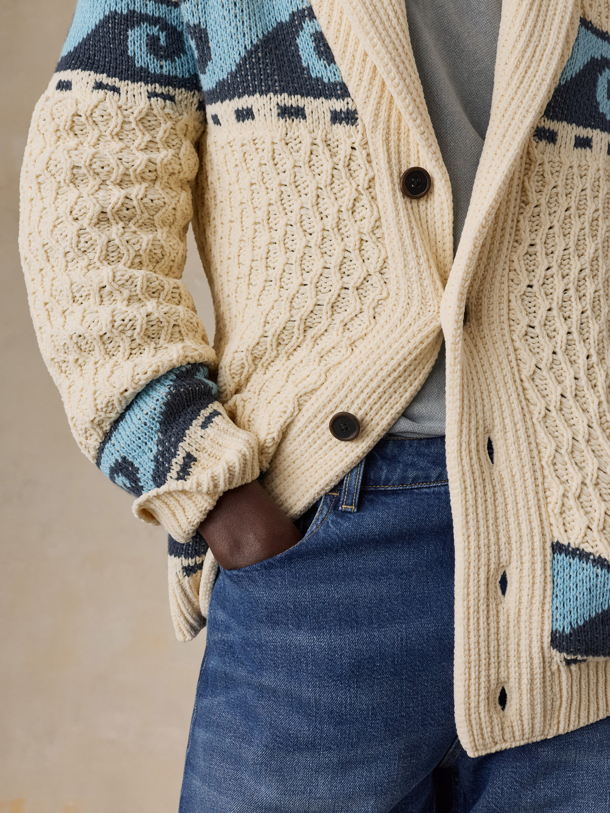 Close up of a man wearing a cream and blue printed cardigan with blue jeans, image 4 of 7