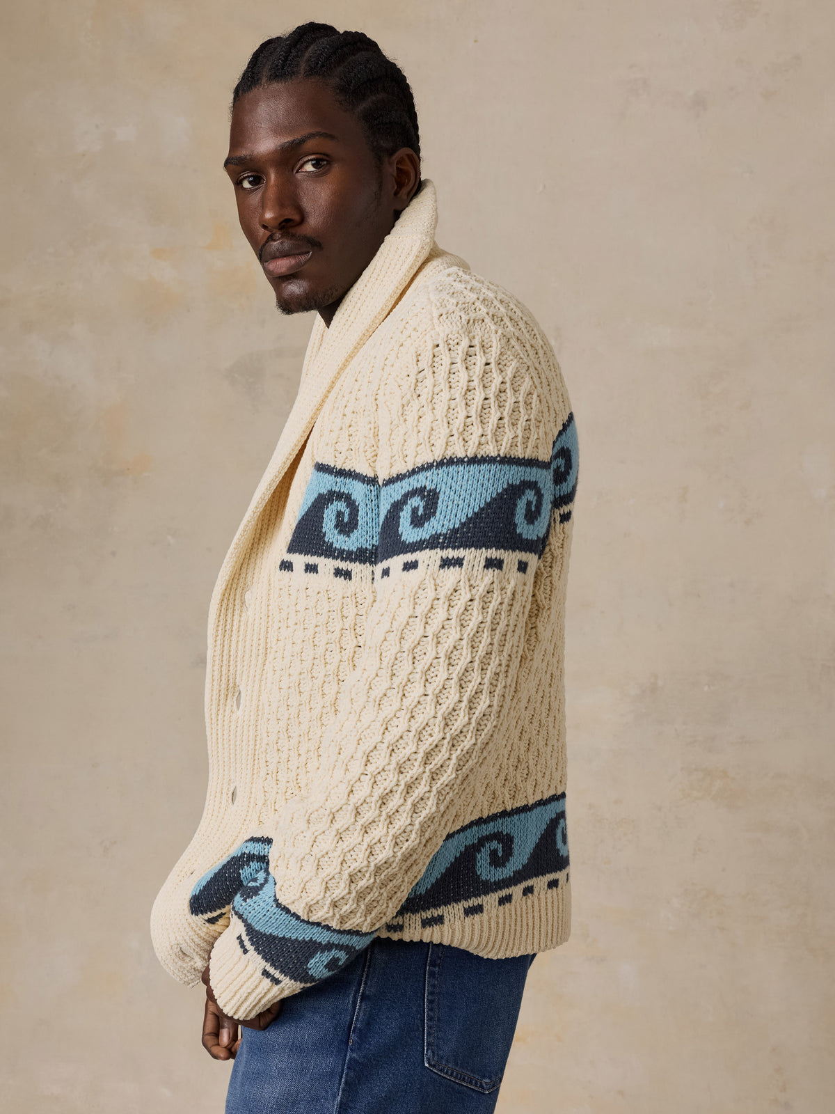 A man wearing a cream and blue printed cardigan with blue jeans, image 5 of 7