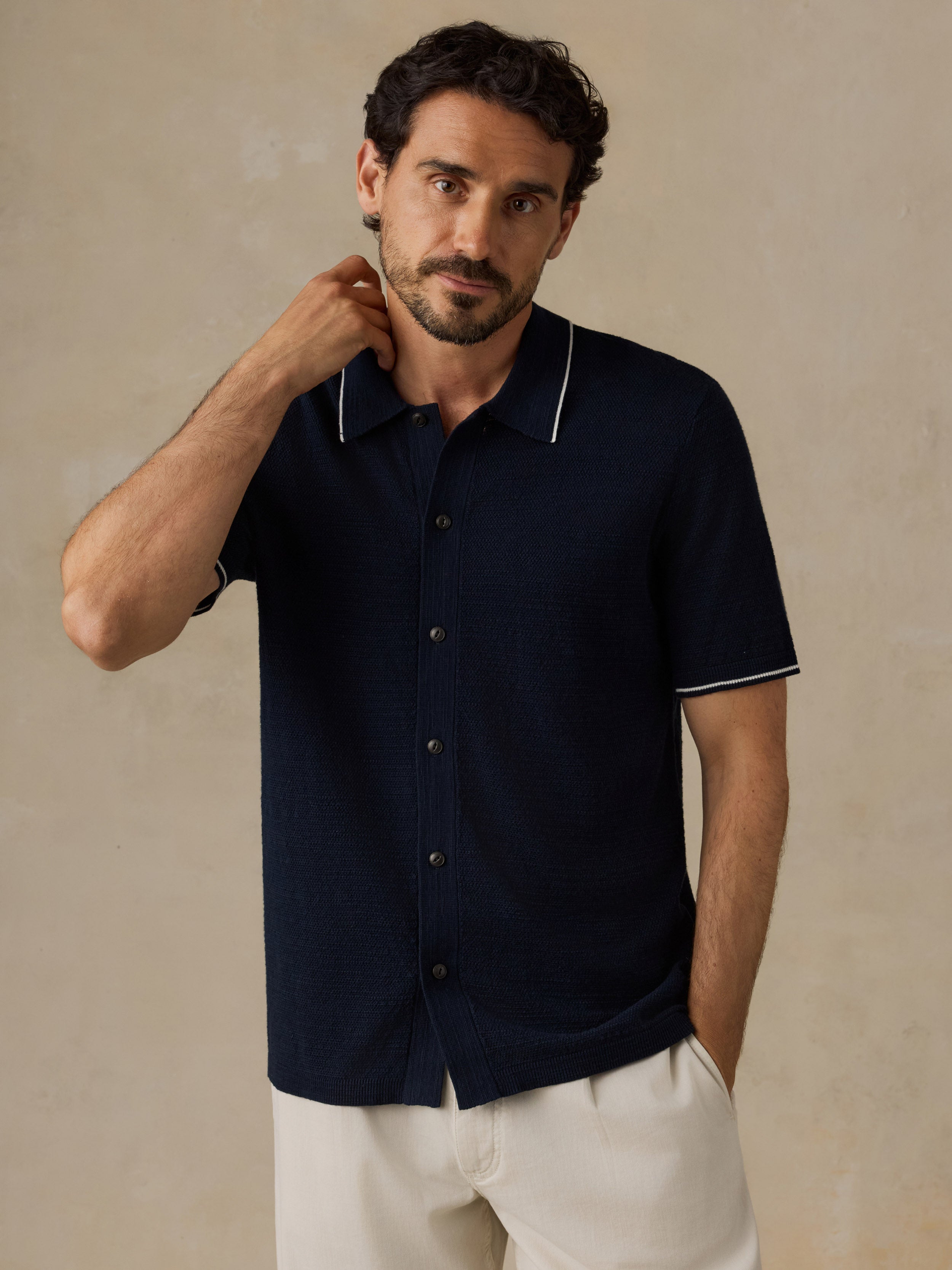 A man wearing a dark navy short-sleeve knit polo shirt with white piping on the collar and sleeve cuffs, paired with off-white trousers, standing against a textured beige background.