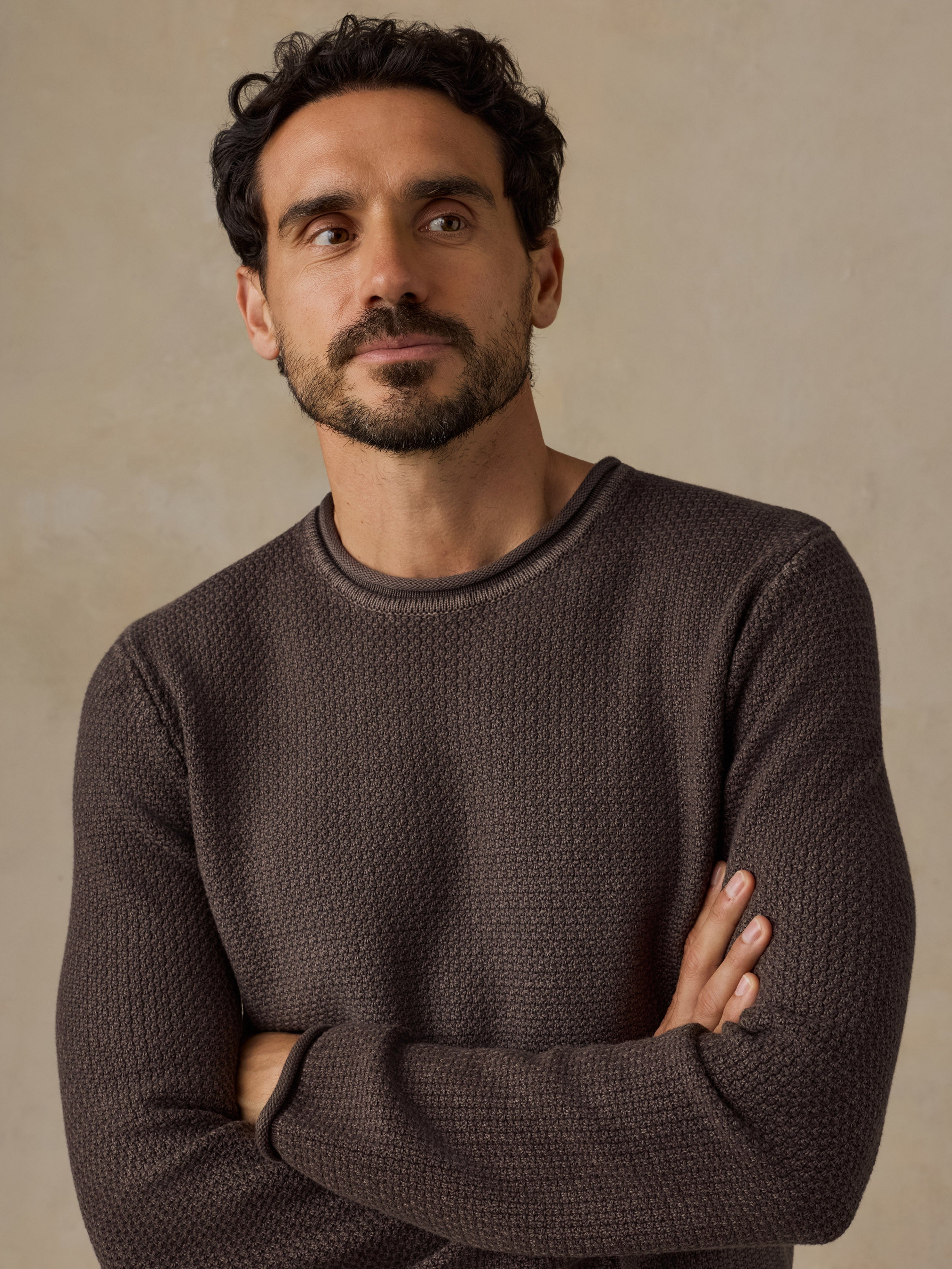 A man with dark curly hair and a beard wearing a textured dark brown crewneck sweater, standing with arms crossed against a warm beige background.