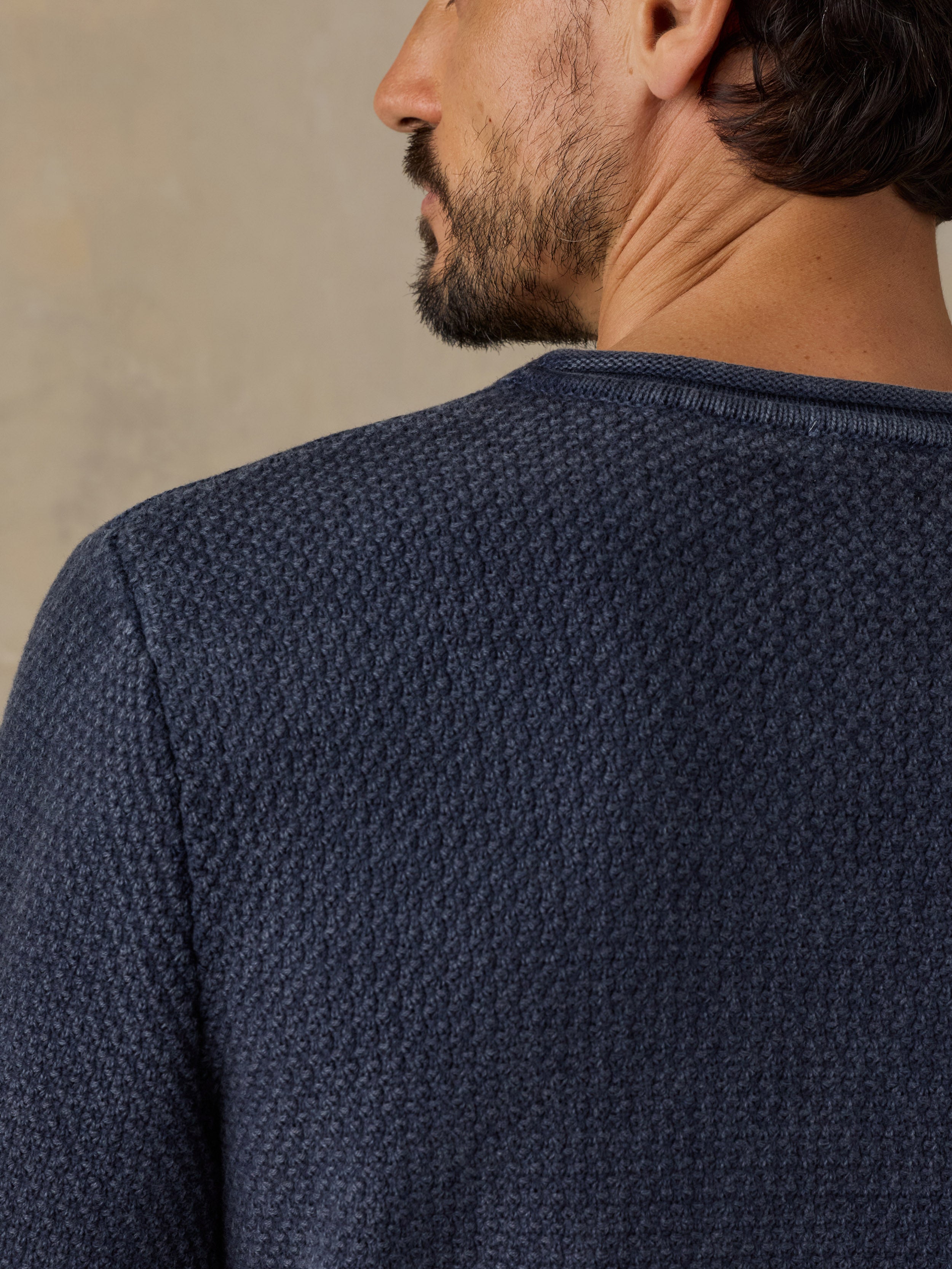 Close-up back view of a man wearing a textured navy blue knit sweater, showing the fine waffle pattern and crew neckline against a warm beige background.