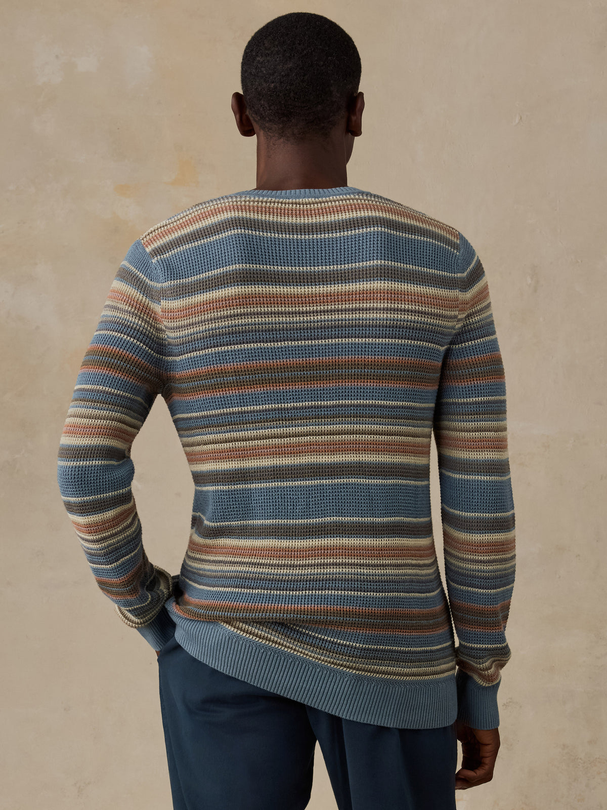 A man wearing a blue, beige, and brown horizontally striped crewneck sweater and dark navy trousers, standing against a beige background., image 3 of 5