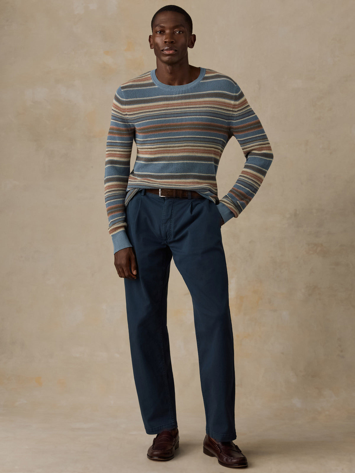 A man wearing a blue, beige, and brown horizontally striped crewneck sweater and dark navy trousers, standing against a beige background., image 4 of 5