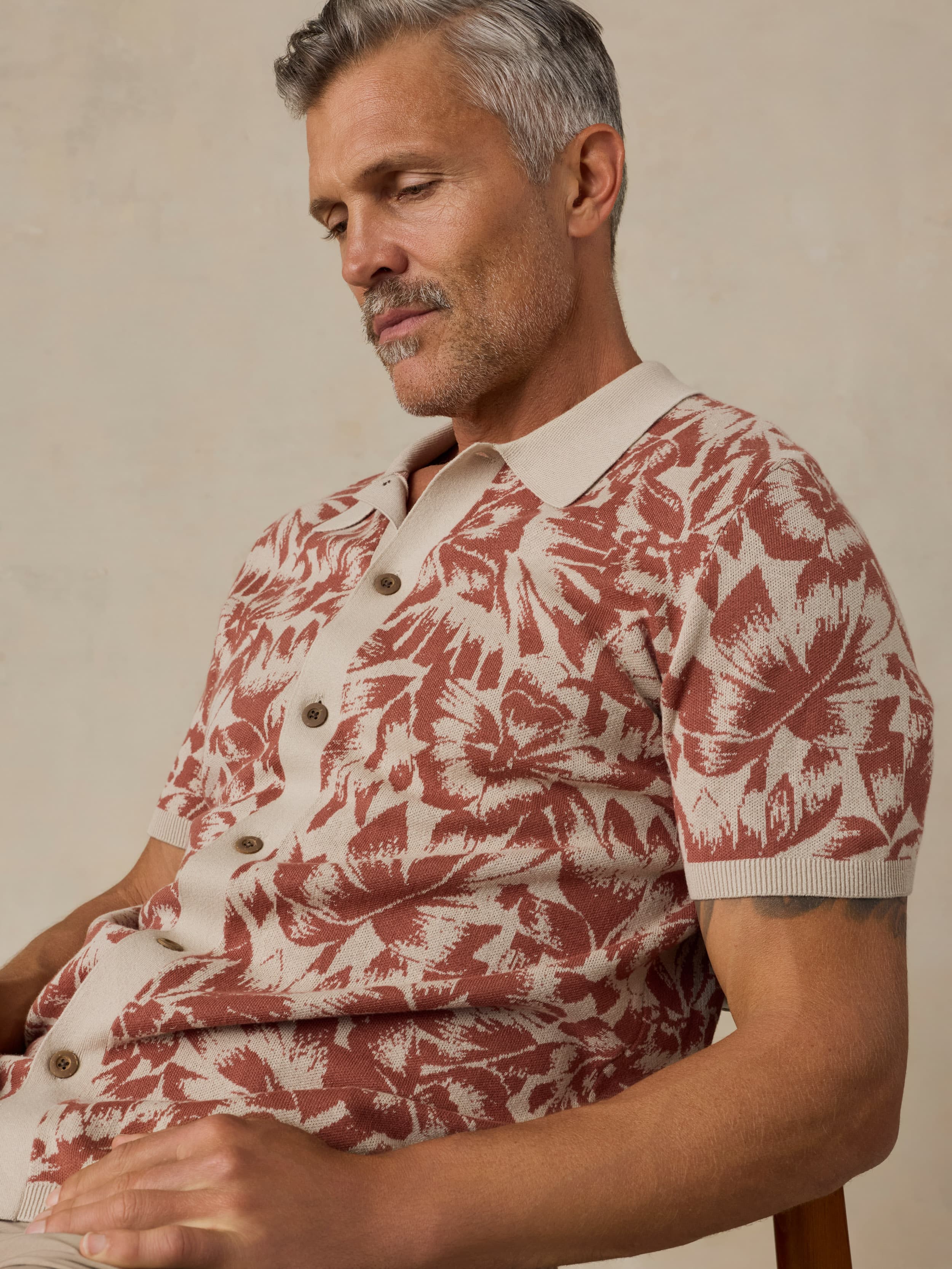 Man wearing a red and white floral patterned shirt sitting on a wooden chair against a beige background