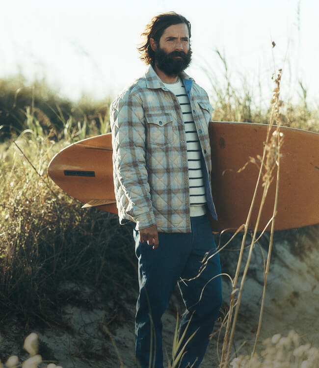Male model carrying surfboard wearing northwoods shirt jacket, lightweight sweater and stretch terry pants