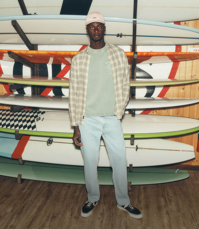 Male model standing infront of surf board rack wearing stretch terry pants, a recycled cotton tee and overshirt paired with a faherty dad cap