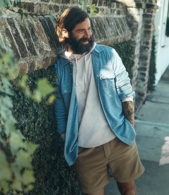 A man leaning against a brick and ivy wall wearing a slub hoodie layered under a legend sweater shirt paired with corduroy shorts