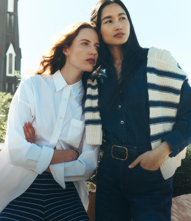 Two women posing on a rooftop, the first wearing a white shirt dress paired with striped knit linen pants, the second wearing a matching indigo shirt and stretch terry pants with a linen sweater draped over her shoulders
