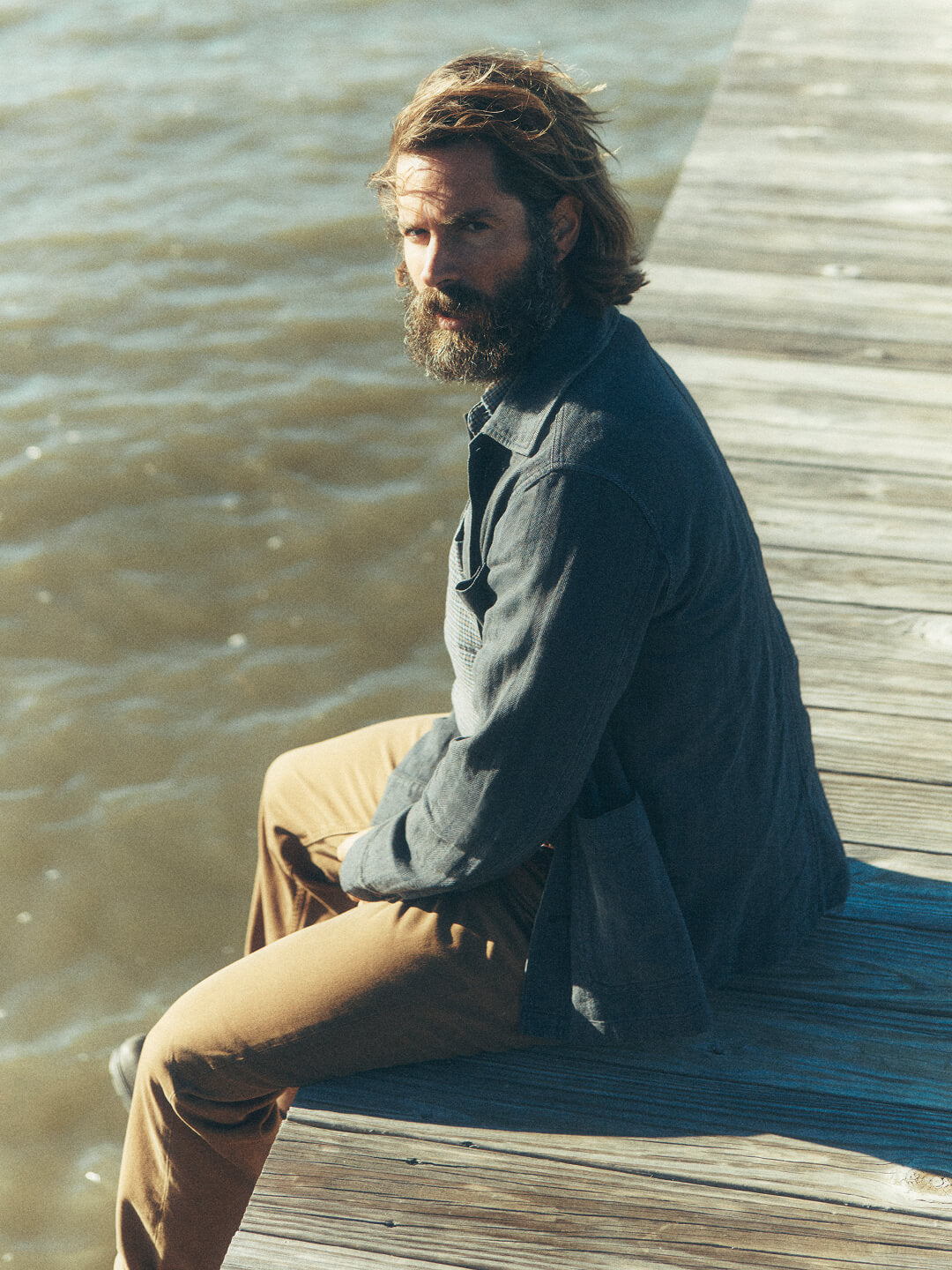 Male model sitting on a dock at sunset wearing an indigo chore coat, a coastline knit shirt and twill pants