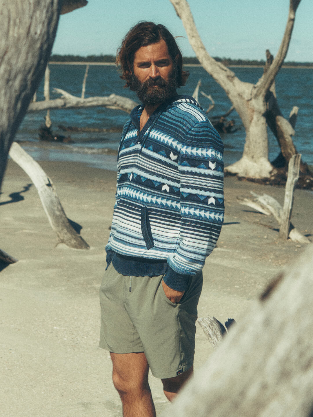 A male model on the beach wearing an indigo hoodie and swim trunks