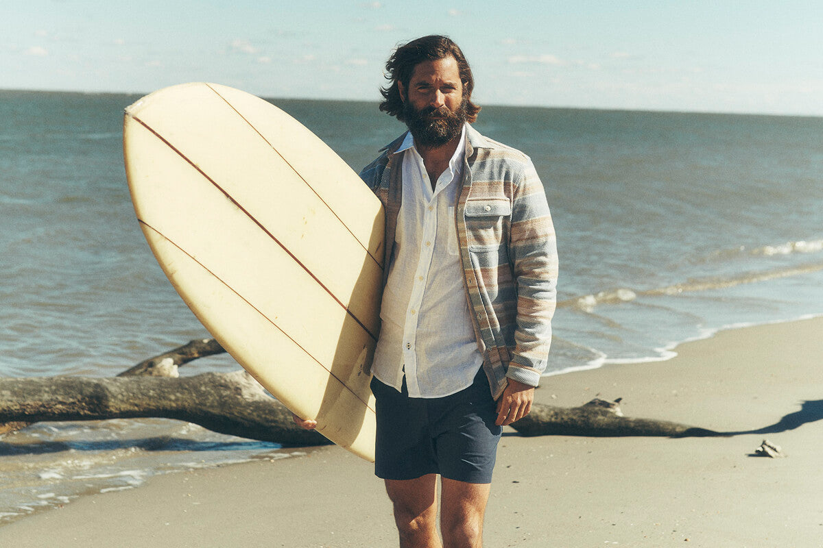 male model carrying a surfboard on the beach, wearing a legend sweater shirt over a white linen shirt paired with all day shorts