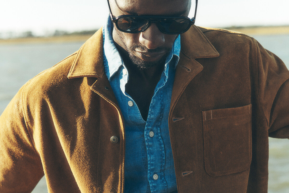 A male model on a dock in Charleston, wearing sunglasses, and a suede chore coat over a chambray shirt and stretch terry pants.