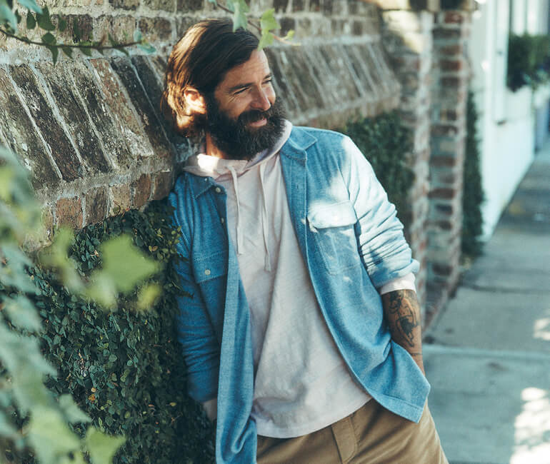 A man leaning against a brick and ivy wall wearing a slub hoodie layered under a legend sweater shirt paired with corduroy shorts