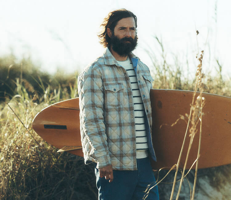 Male model carrying surfboard wearing northwoods shirt jacket, lightweight sweater and stretch terry pants