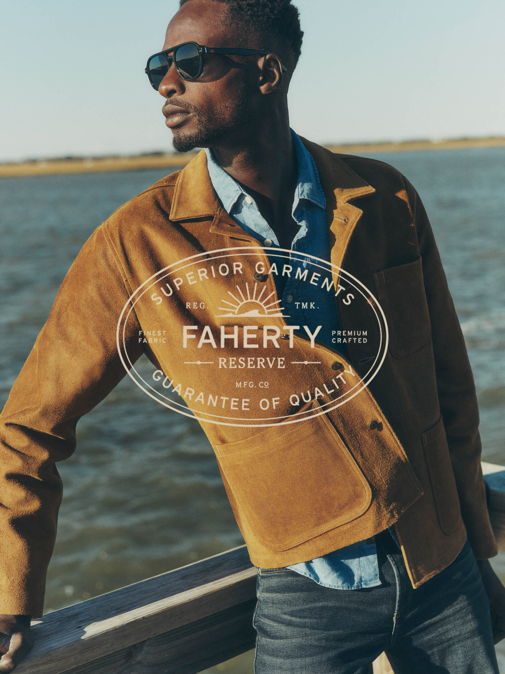 A male model on a dock in Charleston, wearing sunglasses, and a suede chore coat over a chambray shirt and stretch terry pants.