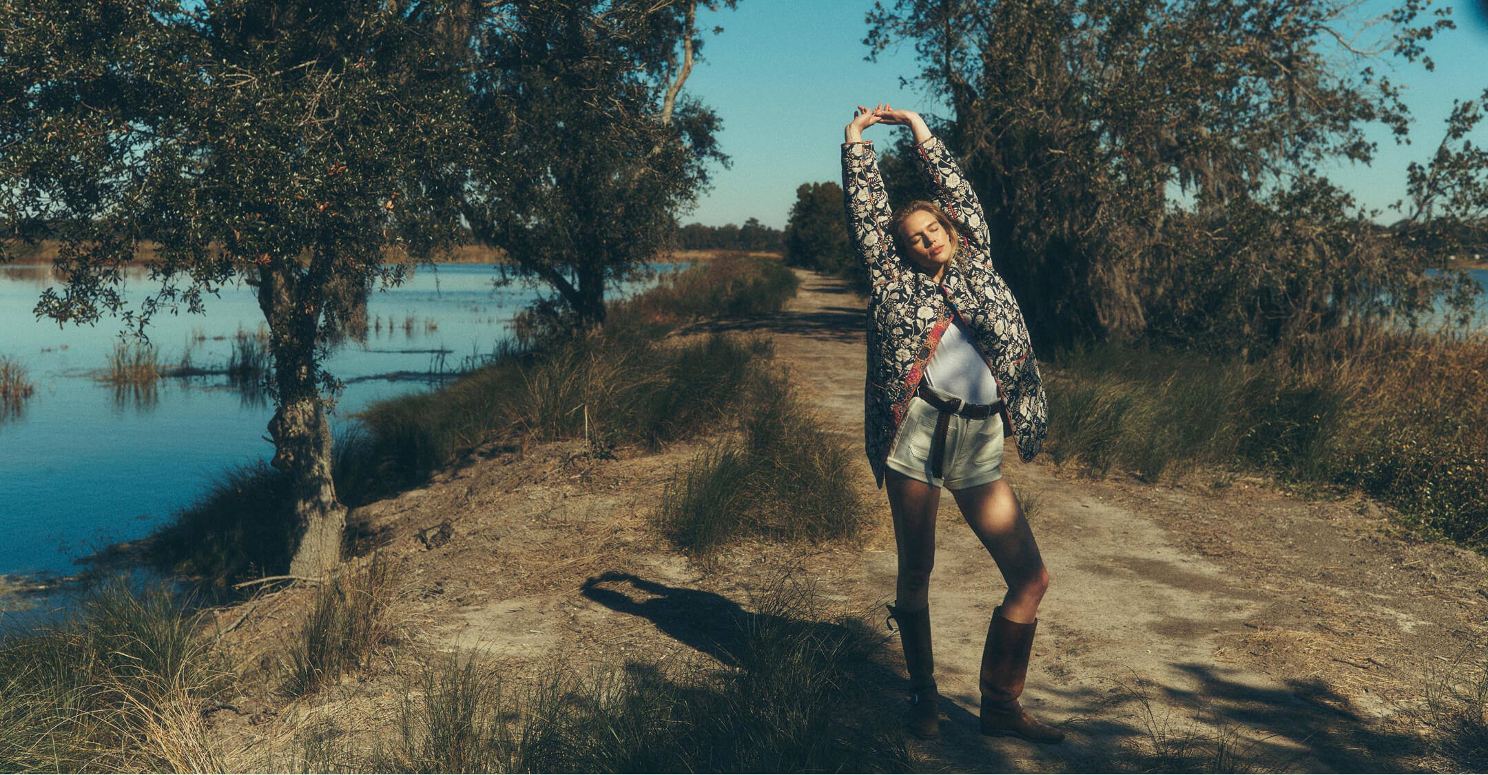 Female model on a dirt pat stretching upwards, wearing a reversible quilted jacket, cotton tee, stretch terry shorts with a braided belt and riding boots.