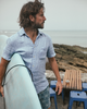 Alex Faherty on the beach with a surfboard, wearing Palma Linen shirt and boardshorts.