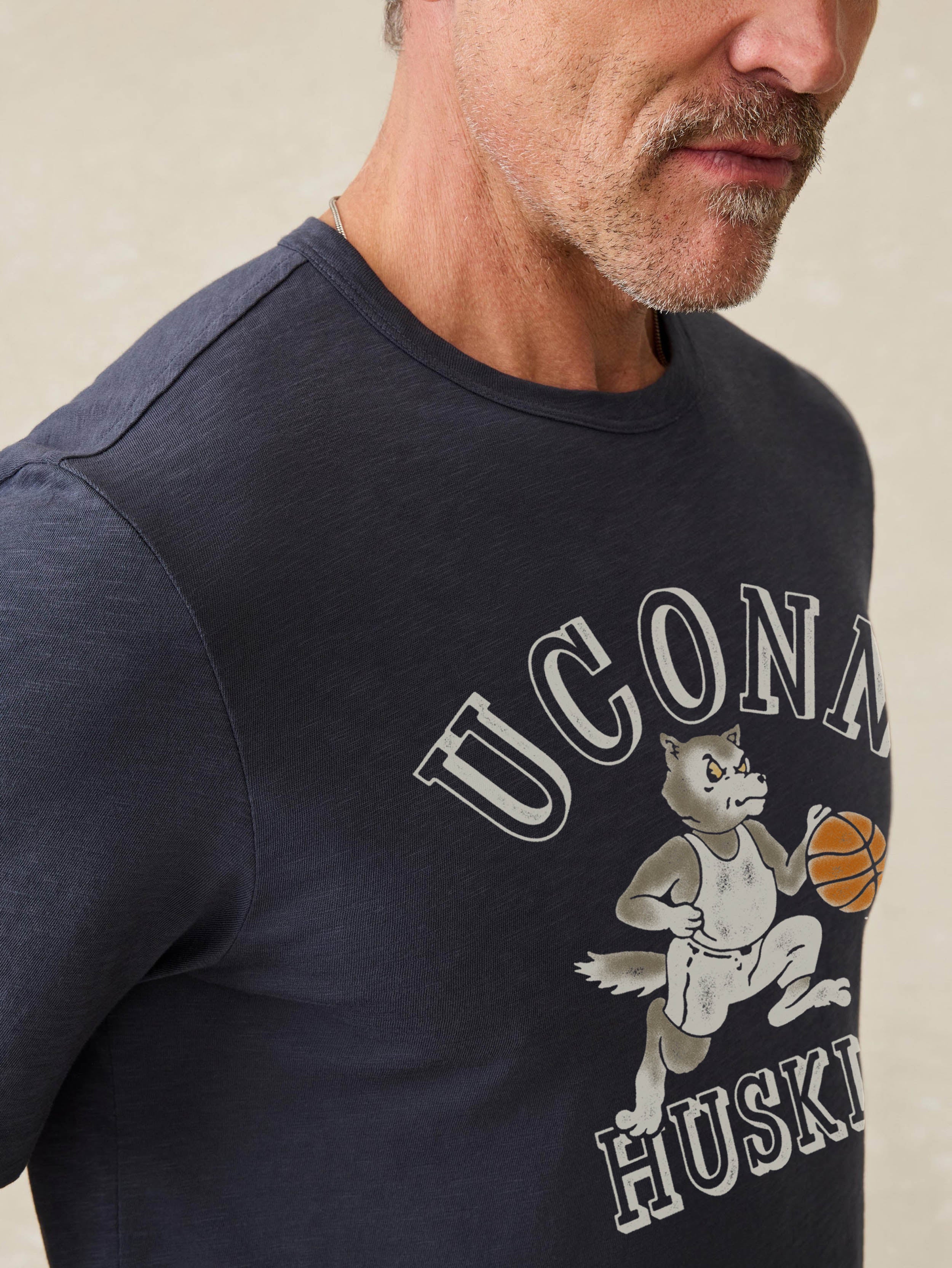 Man wearing a navy blue t-shirt with UConn Huskies graphic on the center front against a beige background