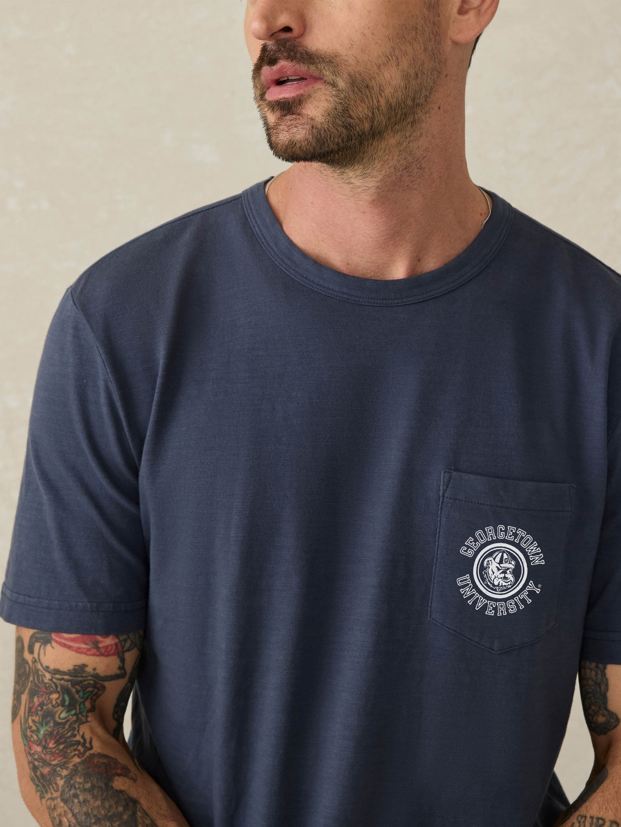 Man wearing a navy blue short sleeve t-shirt with Georgetown graphic on the left chest pocket against a beige background