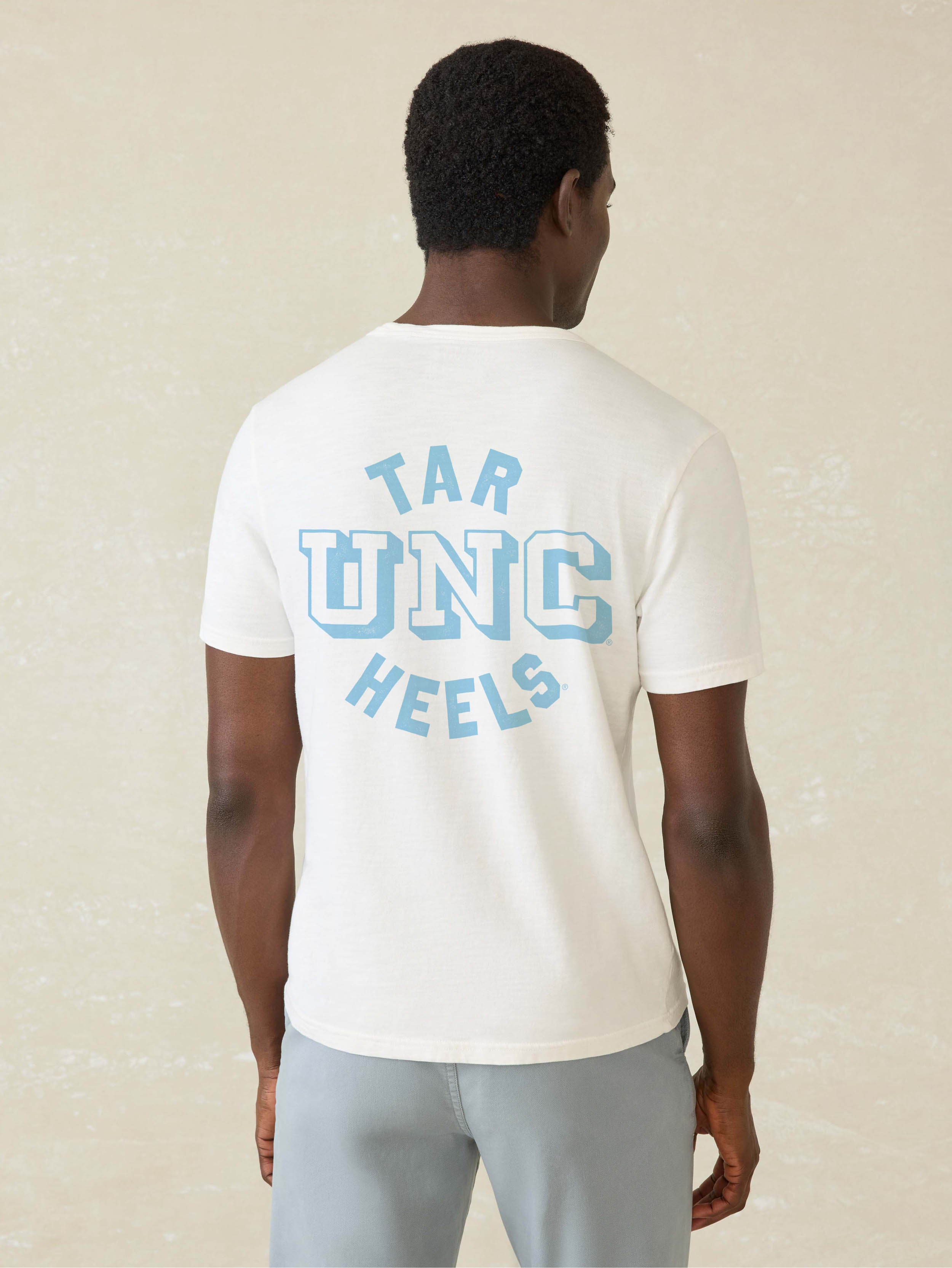Man wearing a white short sleeve t-shirt with UNC graphic at the front chest and the back against a beige background
