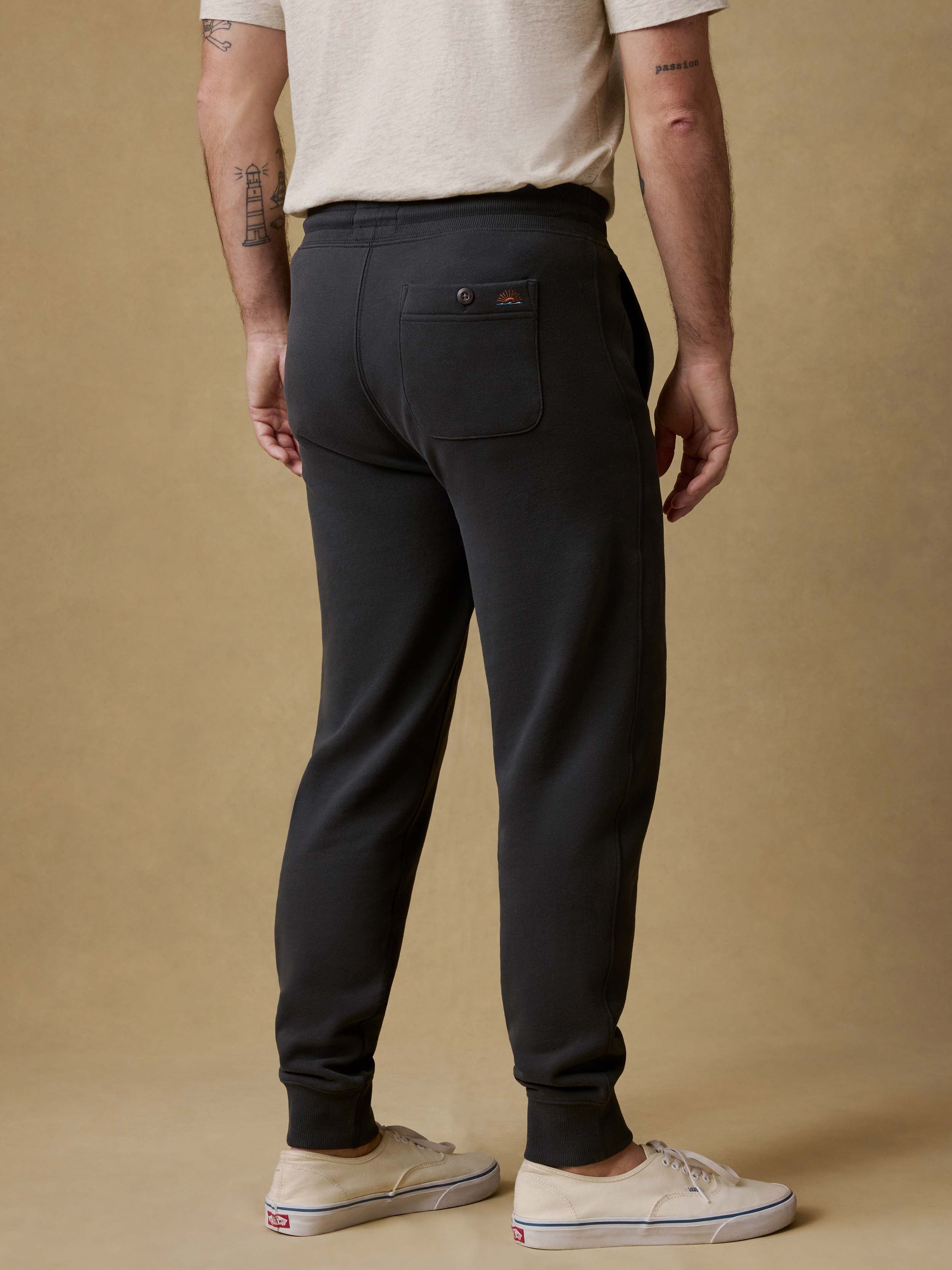 Back view of a man wearing black jogger sweatpants with a white t-shirt and white sneakers on a beige background