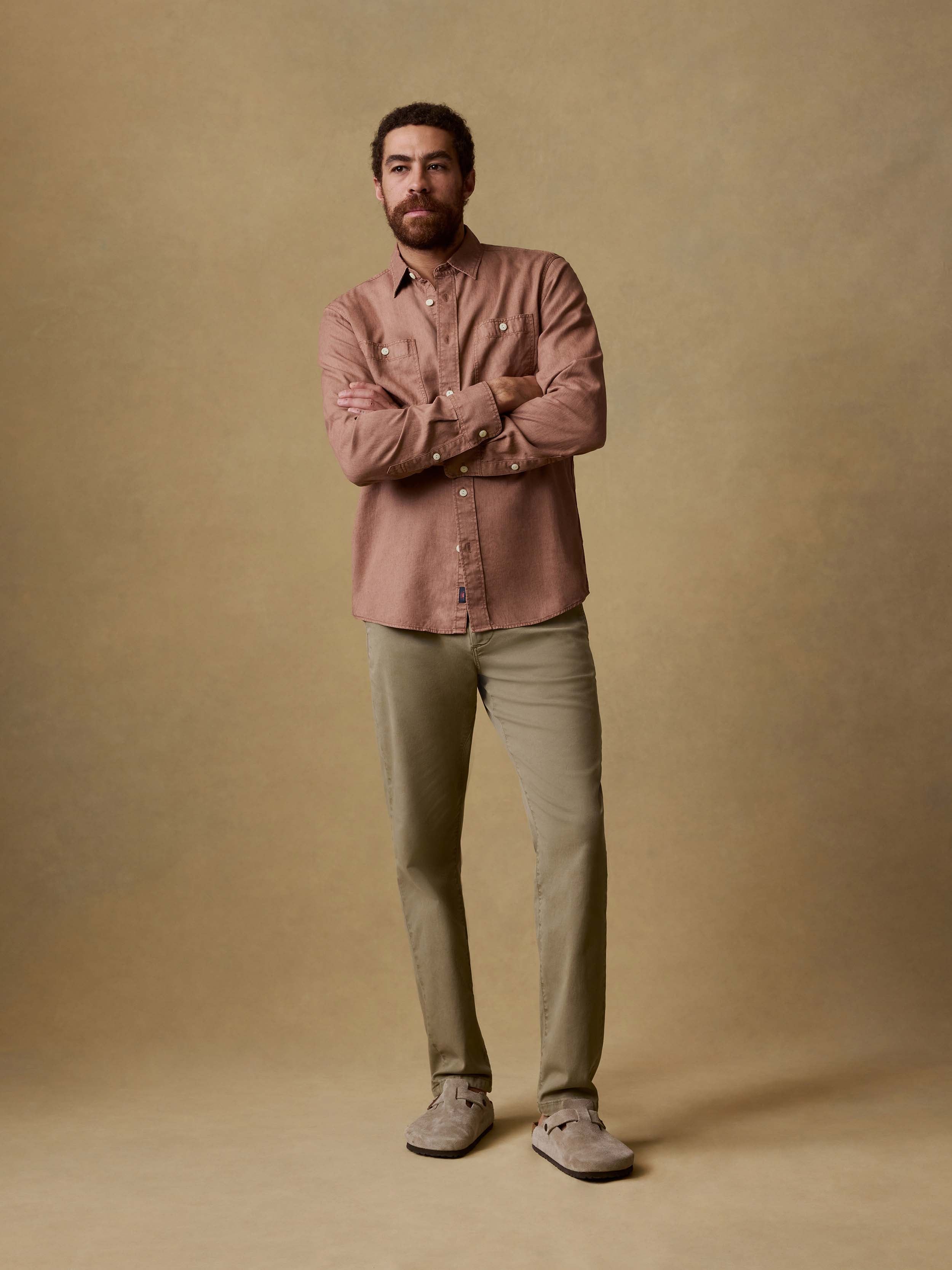 Man is wearing olive pants with a light red button down shirt against a beige background