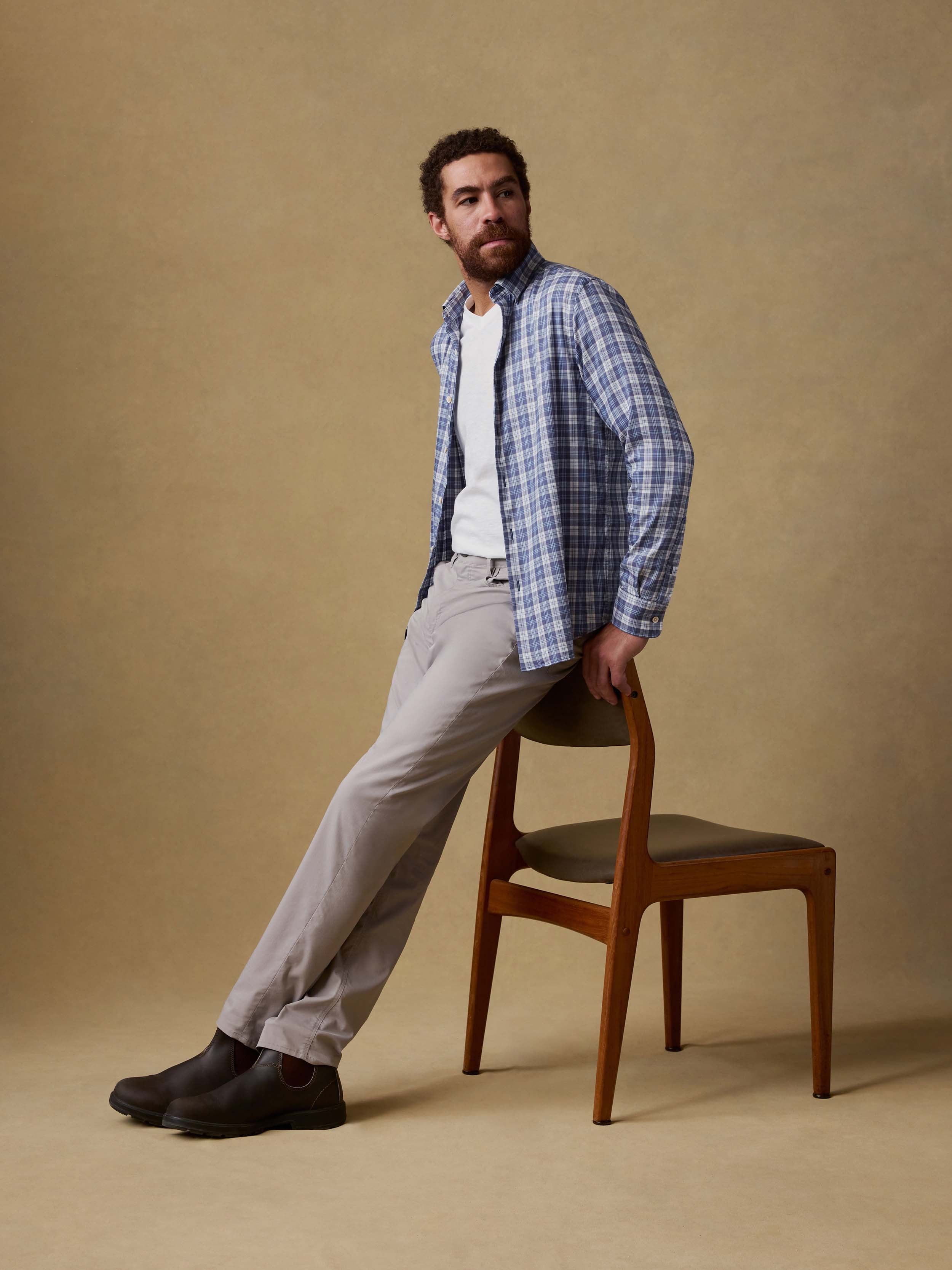Man sitting on a chair wearing a blue plaid shirt, white t-shirt, and light gray pants against a brown background