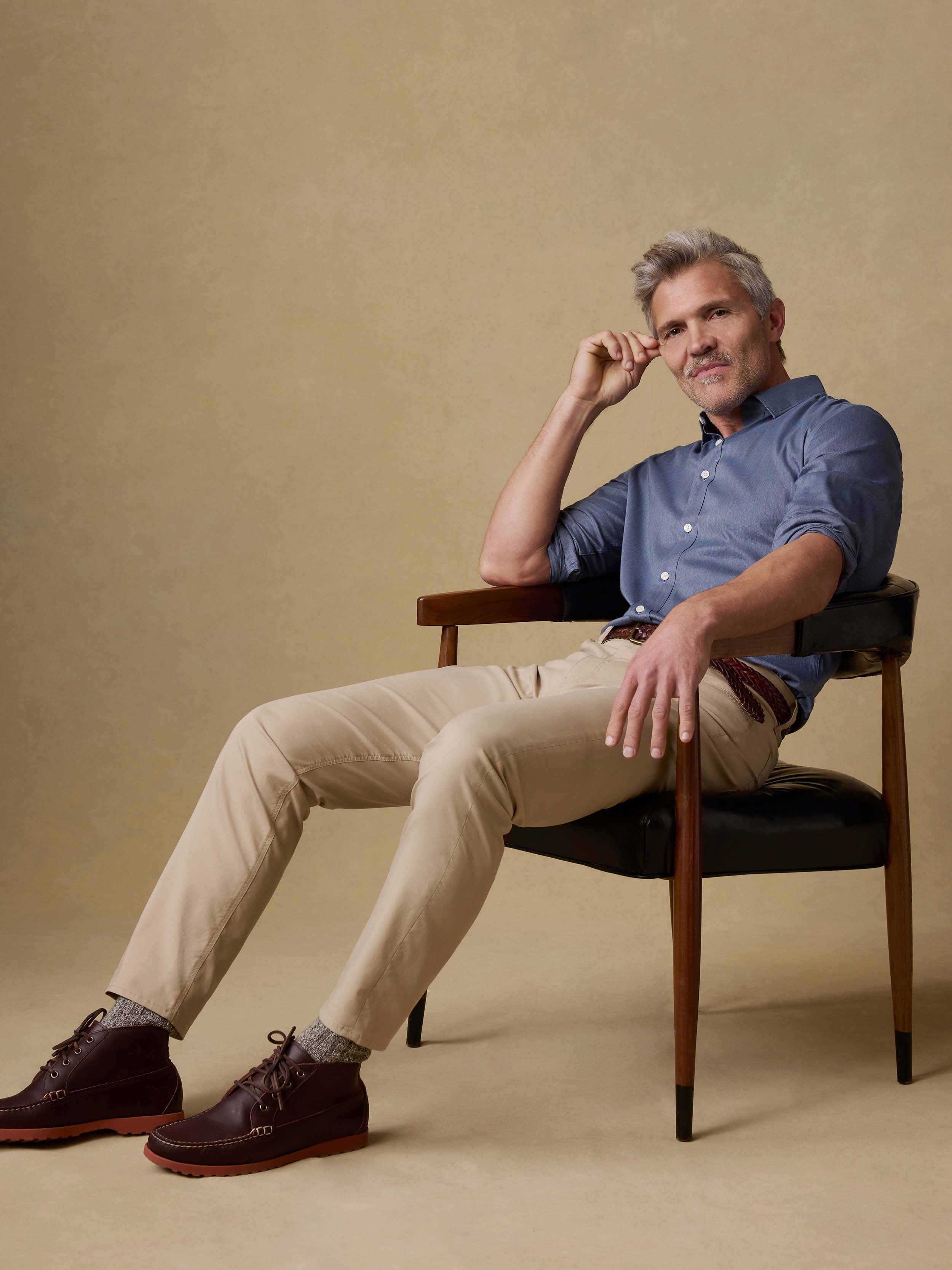 Man sitting on a chair wearing a blue button down shirt and beige pants against a beige background