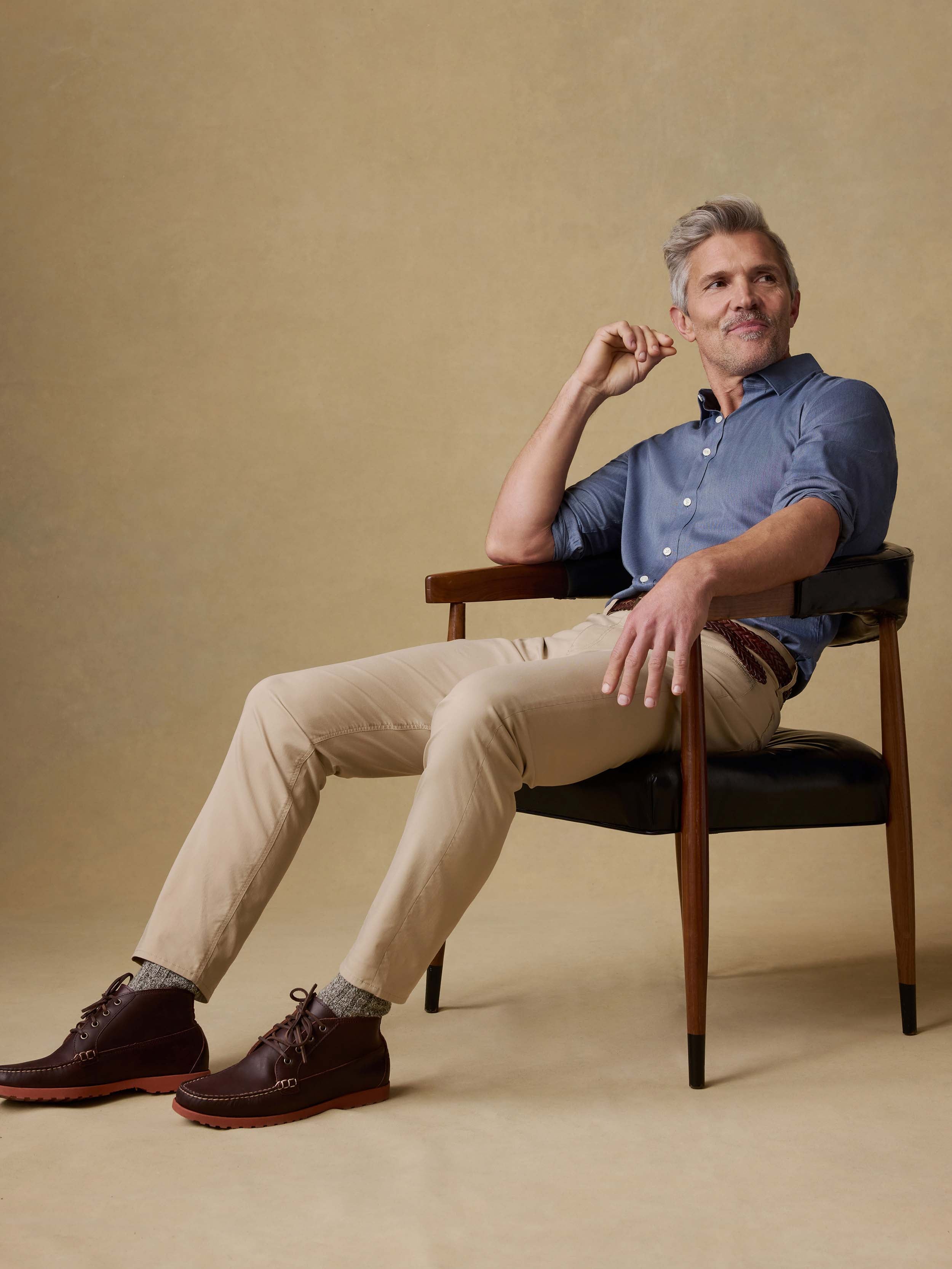 Man sitting on a chair wearing a blue button down shirt and beige pants against a beige background