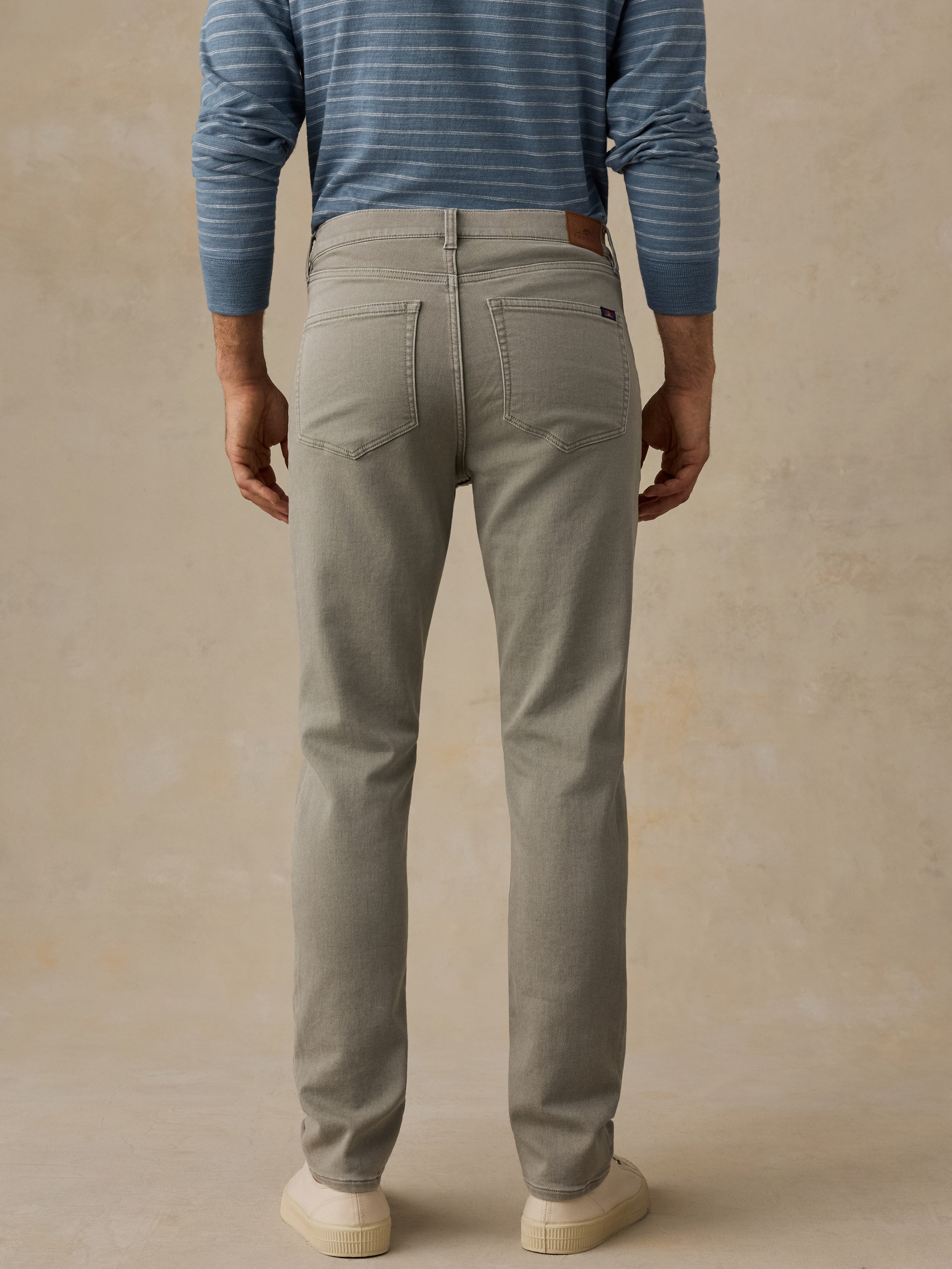 A man viewed from the back wearing light green slim-fit jeans, a blue and white striped long-sleeve hoodie, and cream-colored sneakers, set against a textured beige studio background.