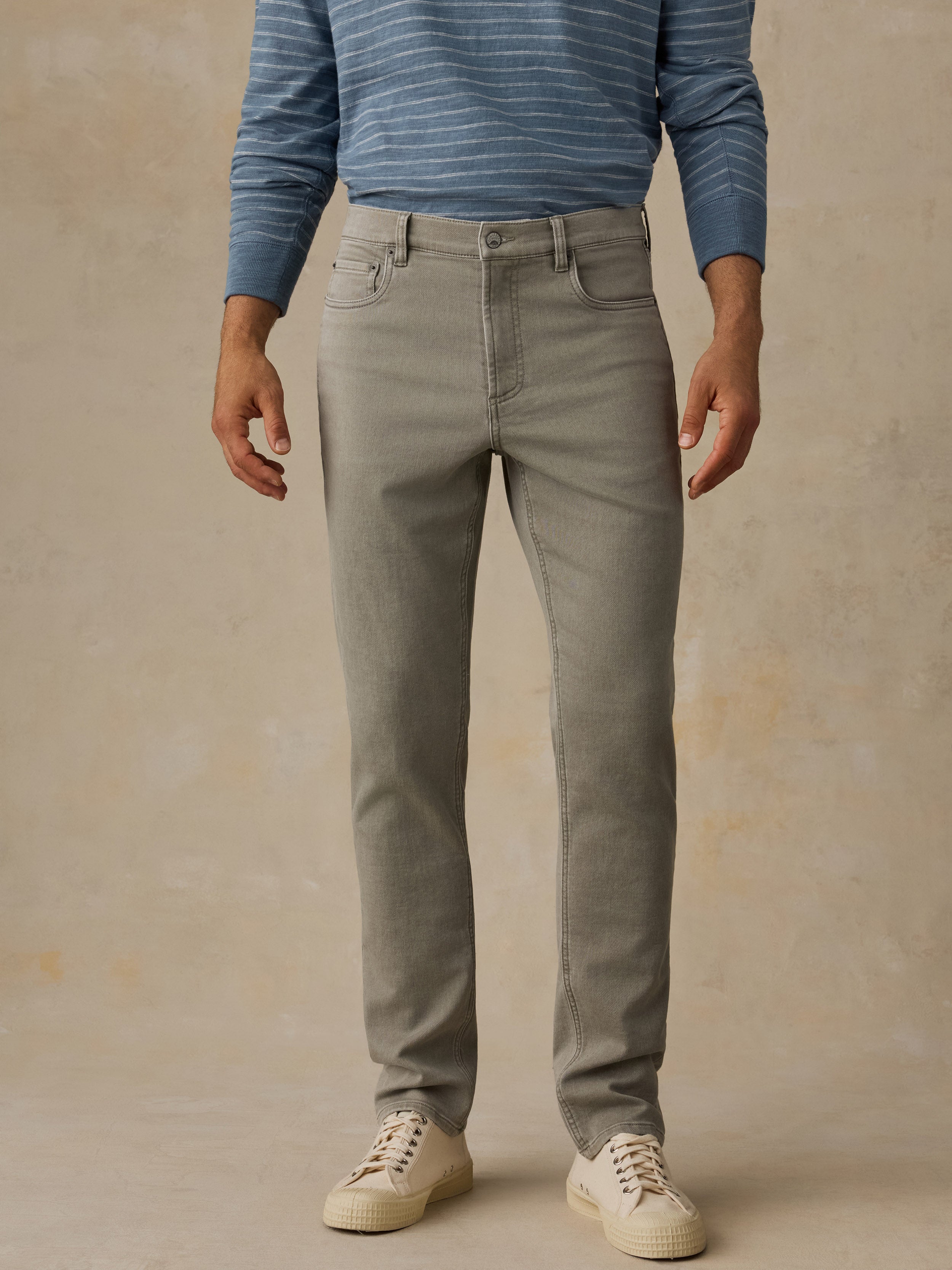A person wearing slim-fit light green jeans, a blue striped long-sleeve shirt, and off-white sneakers, standing against a textured beige studio backdrop.