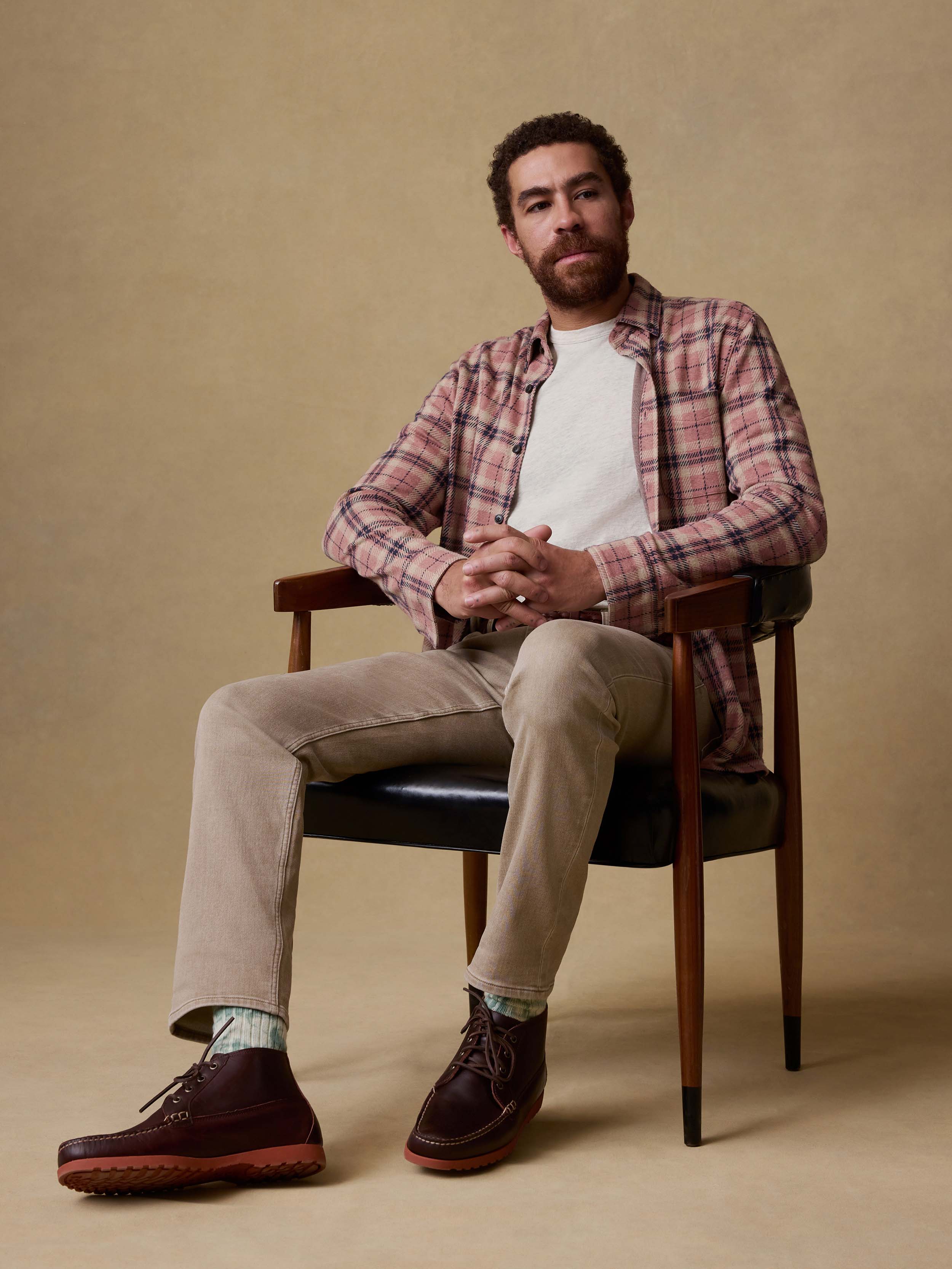 Man sitting on a chair wearing a plaid shirt, white t-shirt, beige pants, and brown shoes against a beige background.