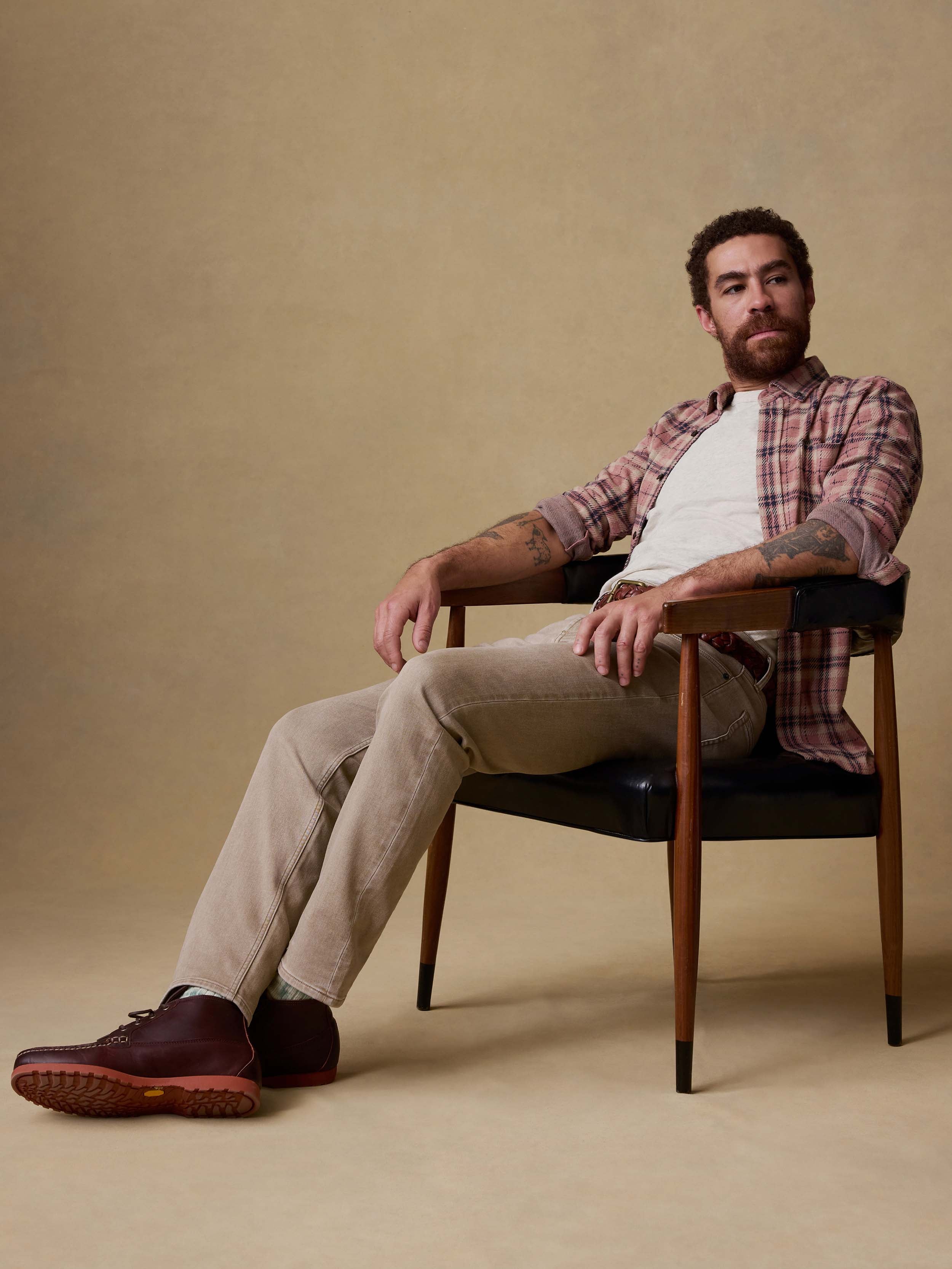 Man sitting on a chair wearing a plaid shirt, beige pants, and brown shoes against a beige background