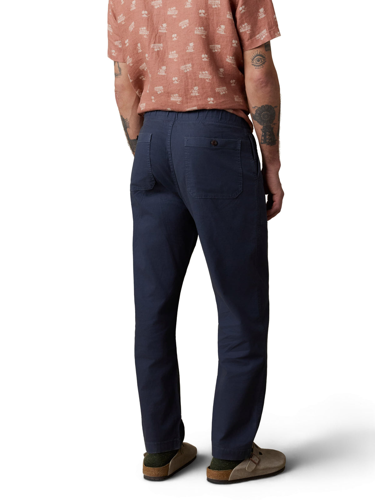 A man viewed from behind wearing an orange button up shirt, blue pants, and birkenstocks, image 3 of 3