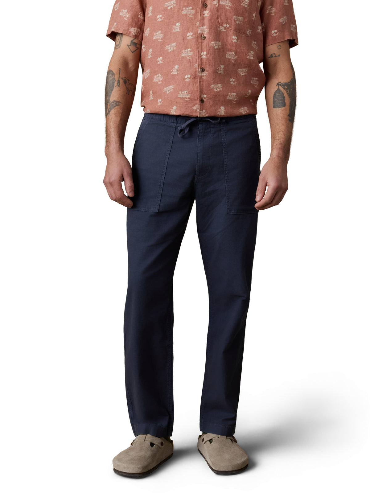 A man wearing an orange button up shirt, blue pants, and birkenstocks, image 2 of 3