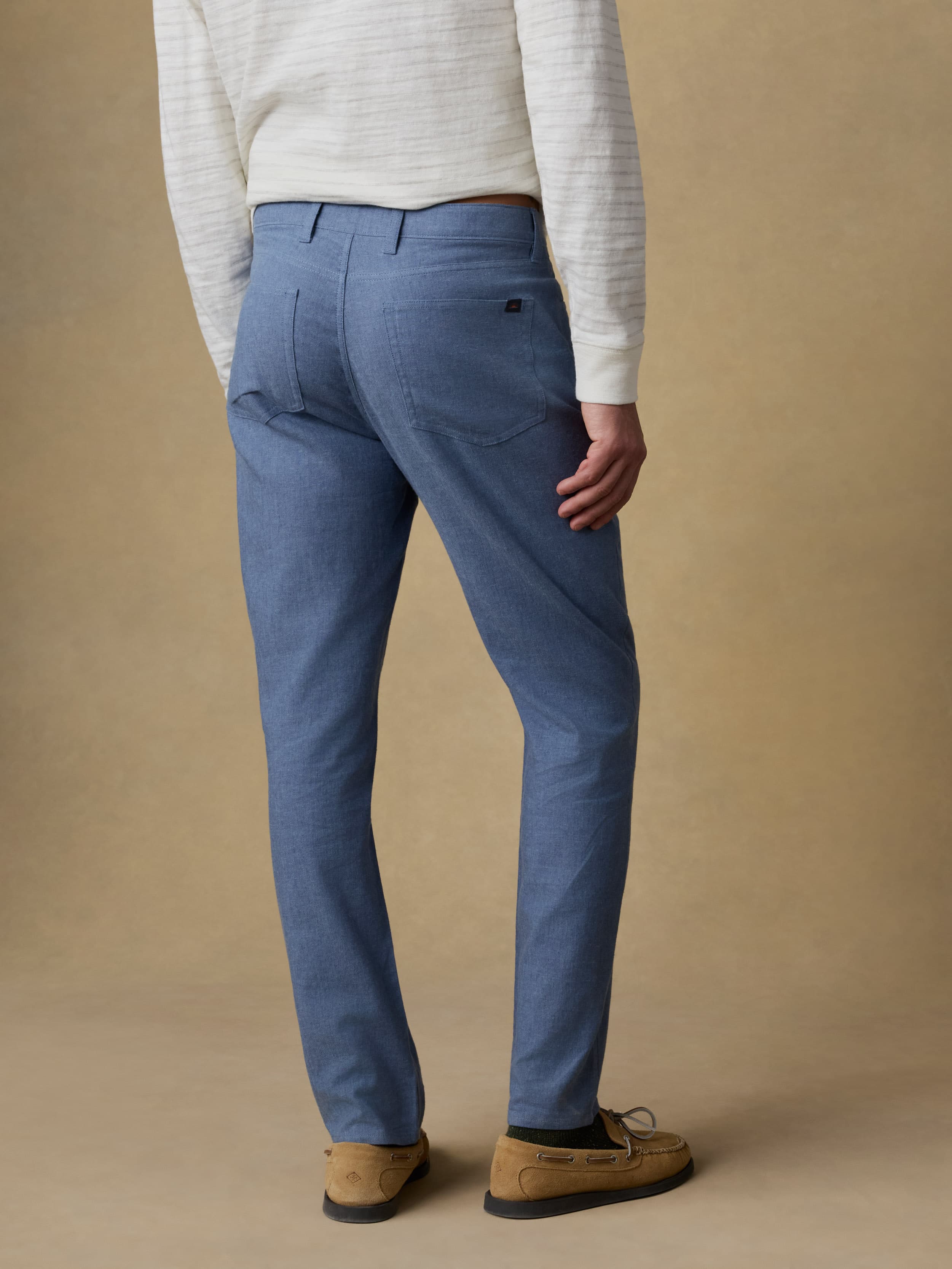 Back view of a man wearing a white and grey striped long sleeve t-shirt with blue linen pants against a beige background