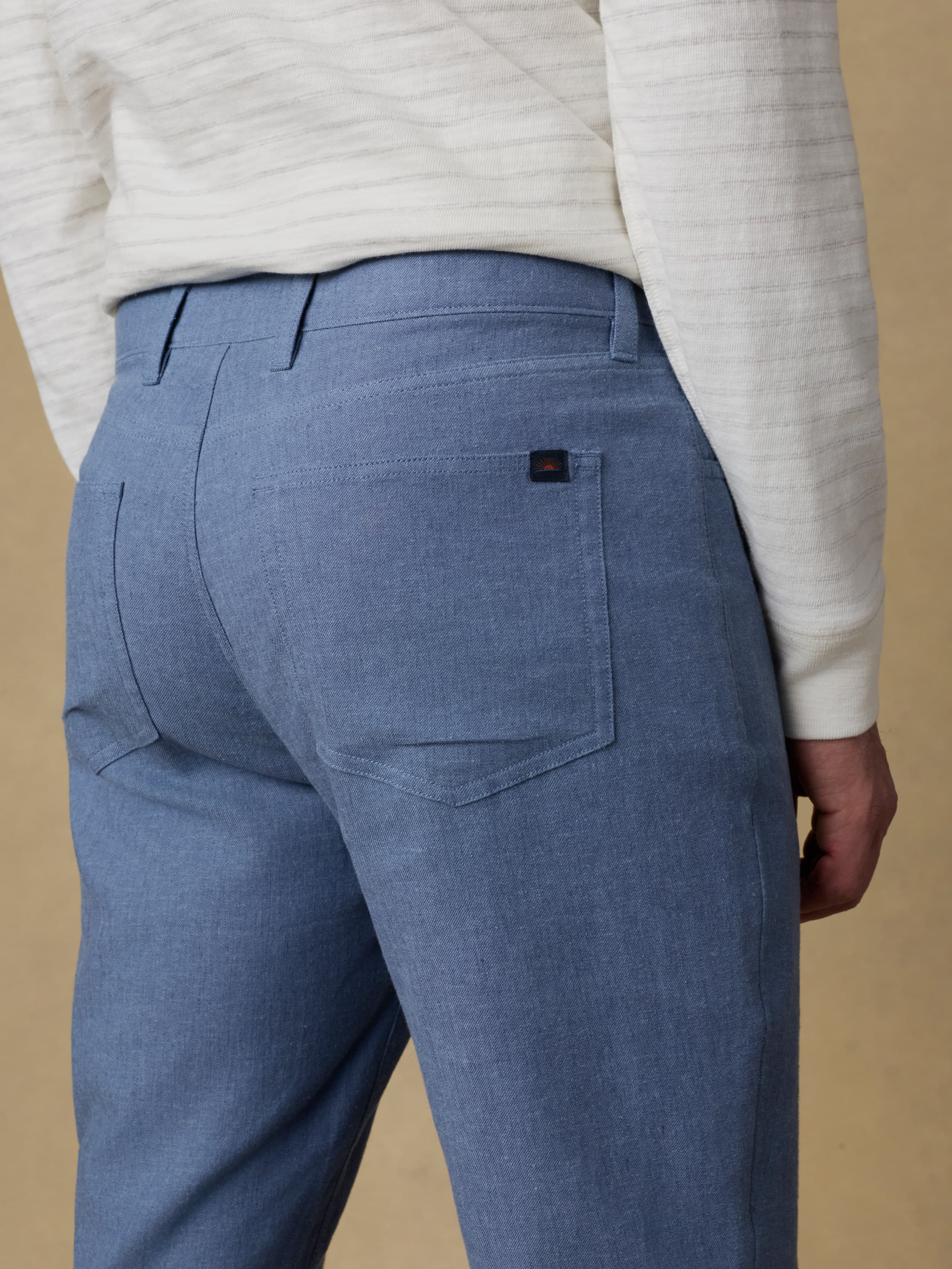 Detail back view of a man wearing a white and grey striped long sleeve t-shirt with blue linen pants against a beige background