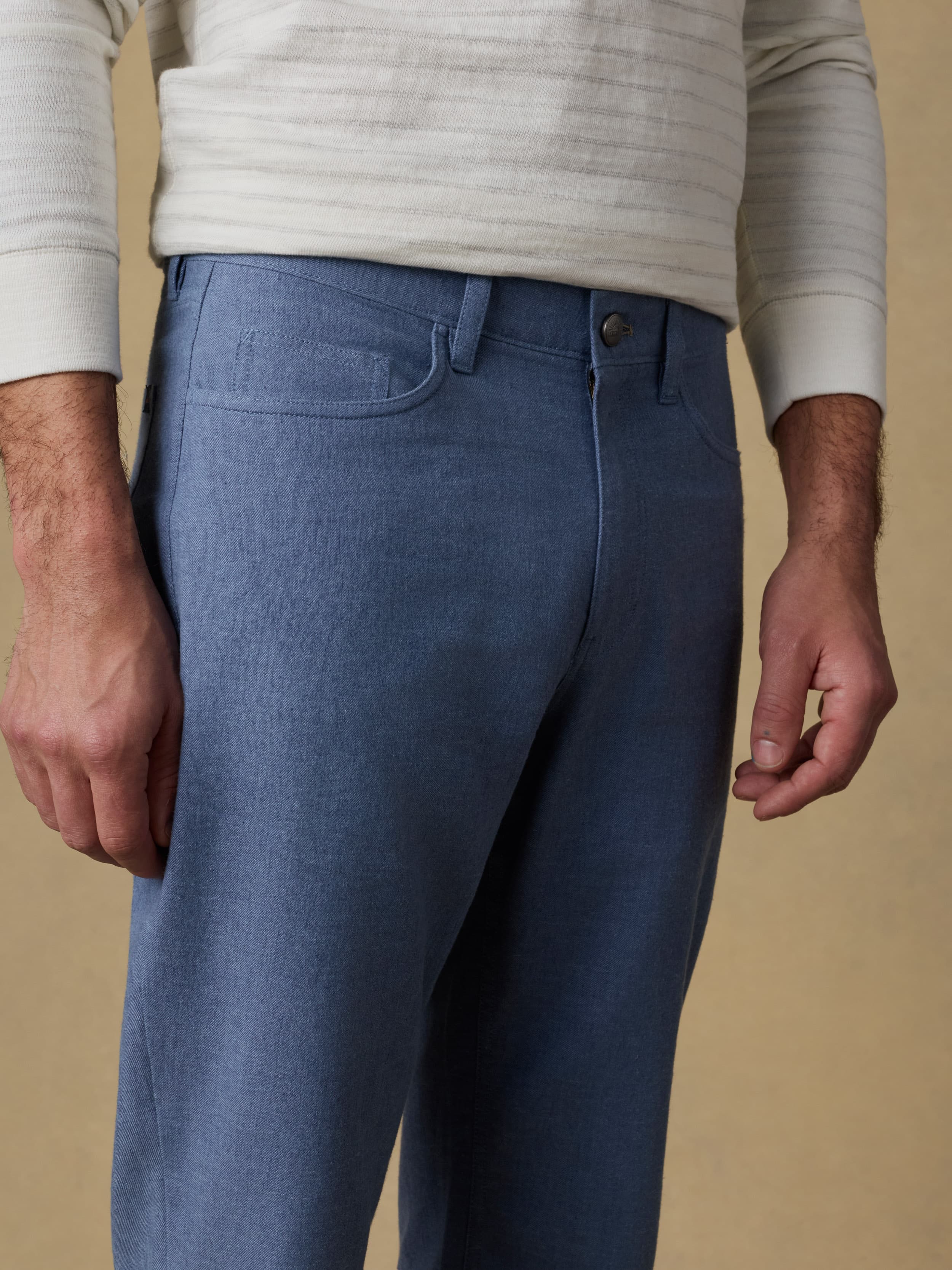 Detail view of a man wearing a white and grey striped long sleeve t-shirt with blue linen pants against a beige background