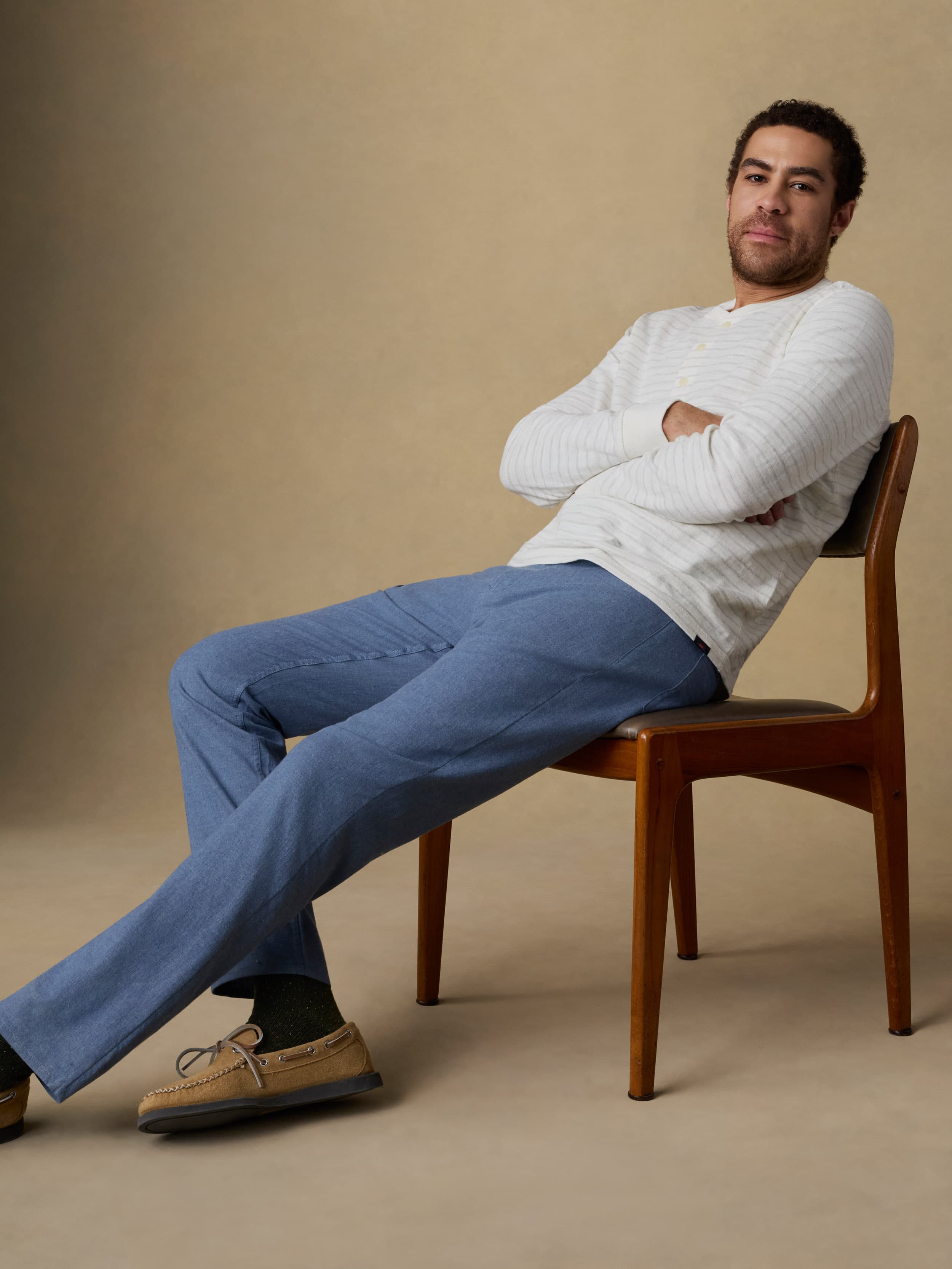 Man sitting on a wooden chair wearing a white and grey striped long sleeve t-shirt with blue linen pants against a beige background