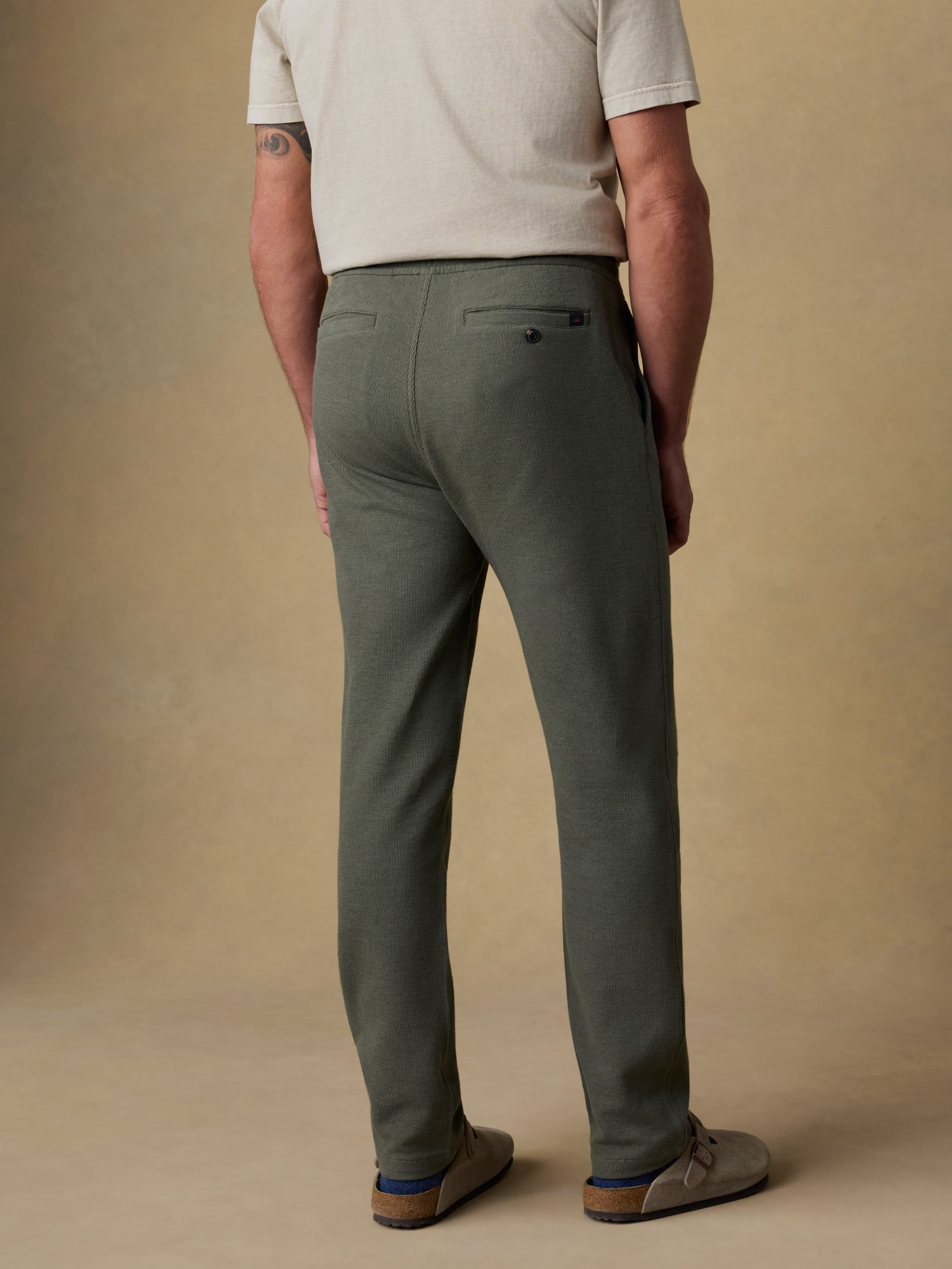 Back view of a man wearing a beige t-shirt with green sweatpants against a beige background