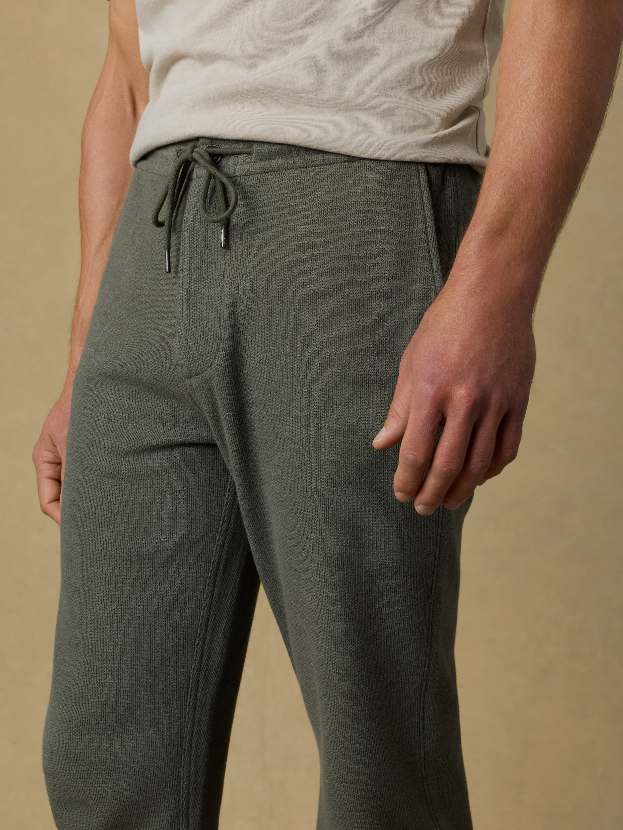 Detail shot of a man wearing a beige t-shirt with green sweatpants against a beige background