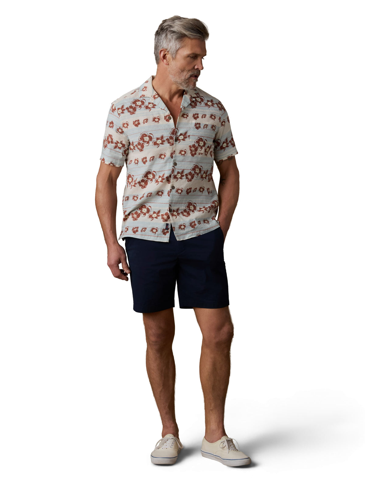 A man wearing a printed button up shirt, navy shorts, and white sneakers, image 1 of 3